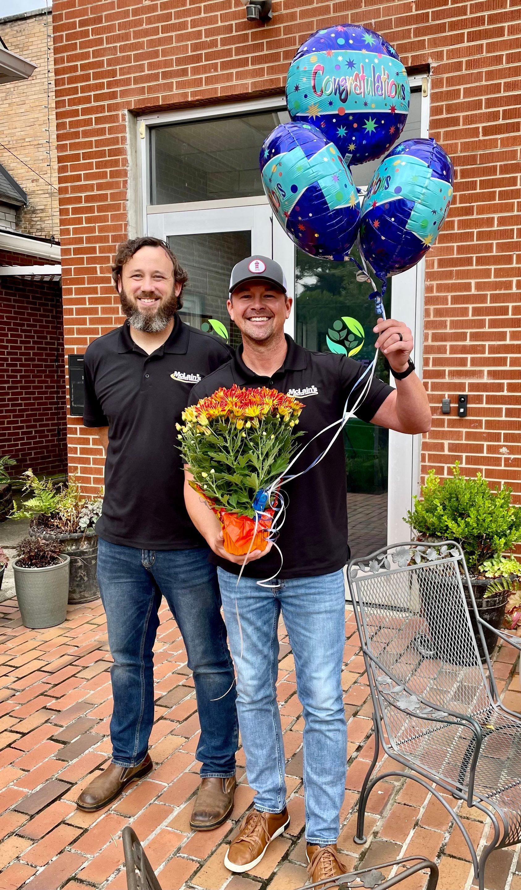 Two men are standing next to each other holding balloons and flowers.