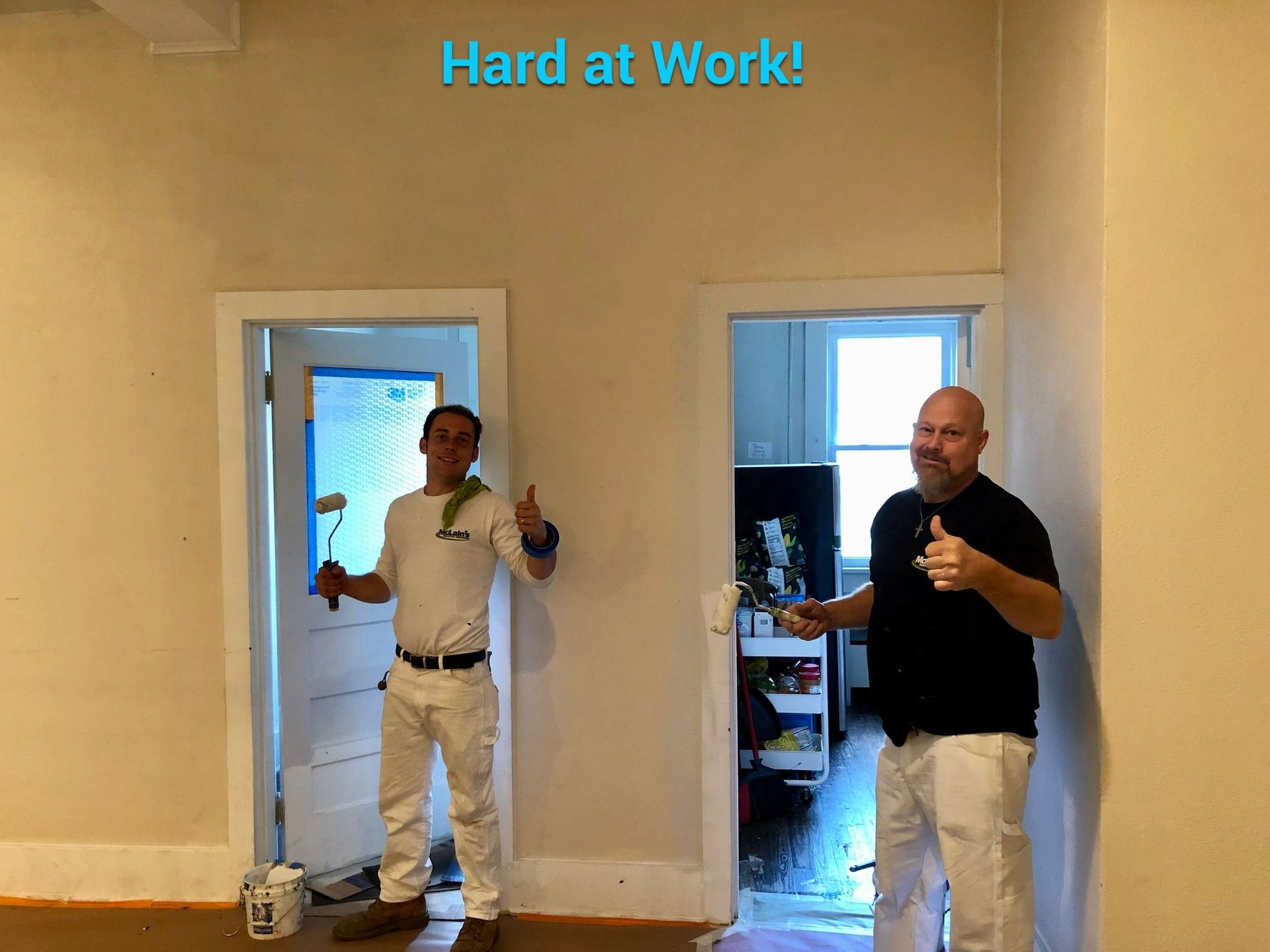 Two men are standing in a room holding paint rollers and giving a thumbs up.
