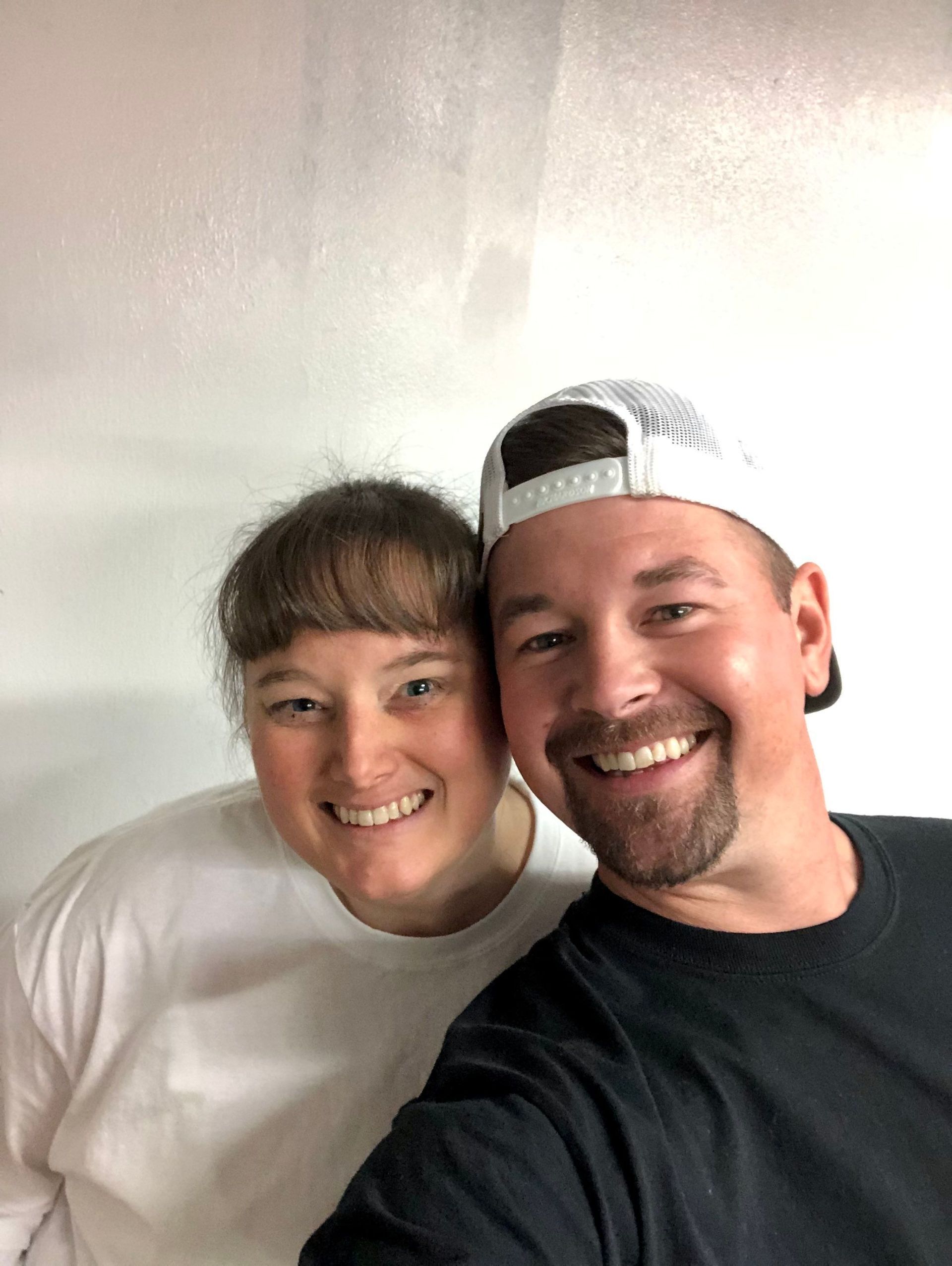 A man and a woman are posing for a selfie in front of a white wall.