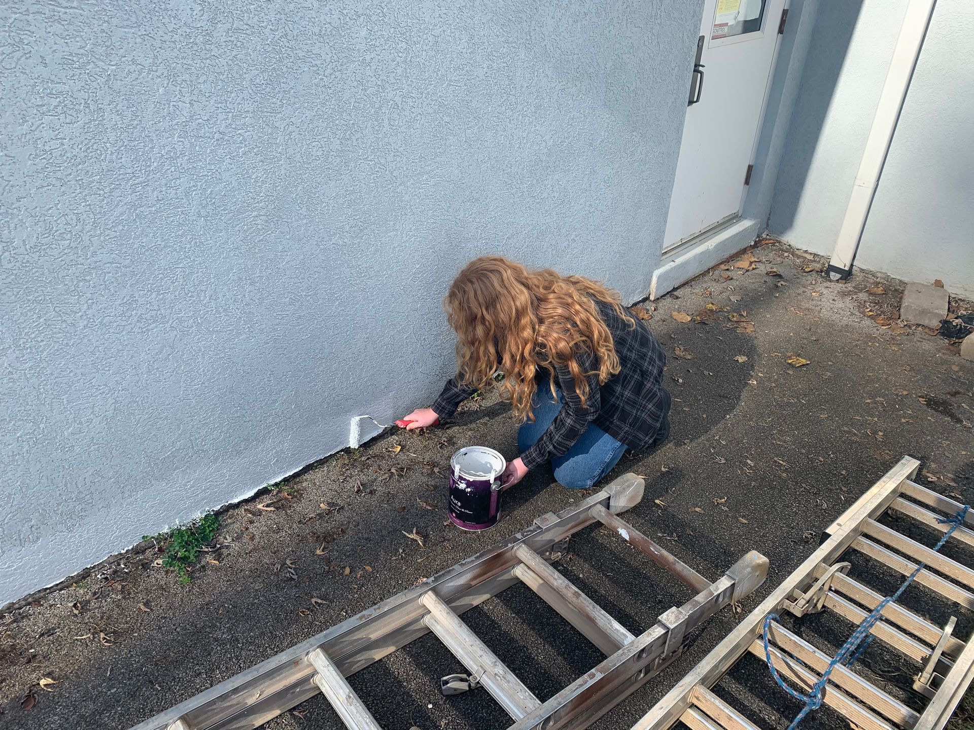 A woman is kneeling on the ground painting a wall next to a ladder.