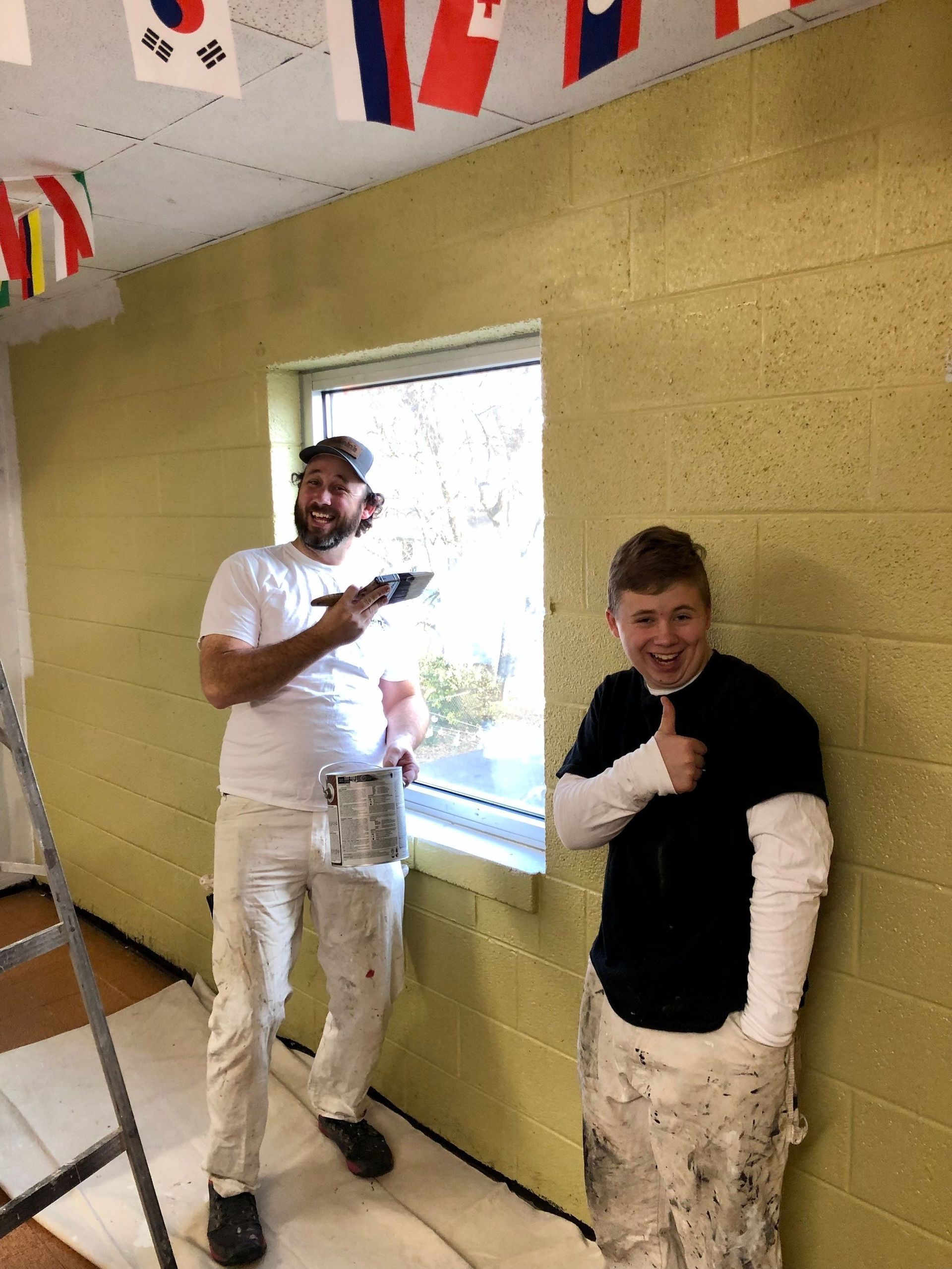 Two men are painting a wall and one is giving a thumbs up.
