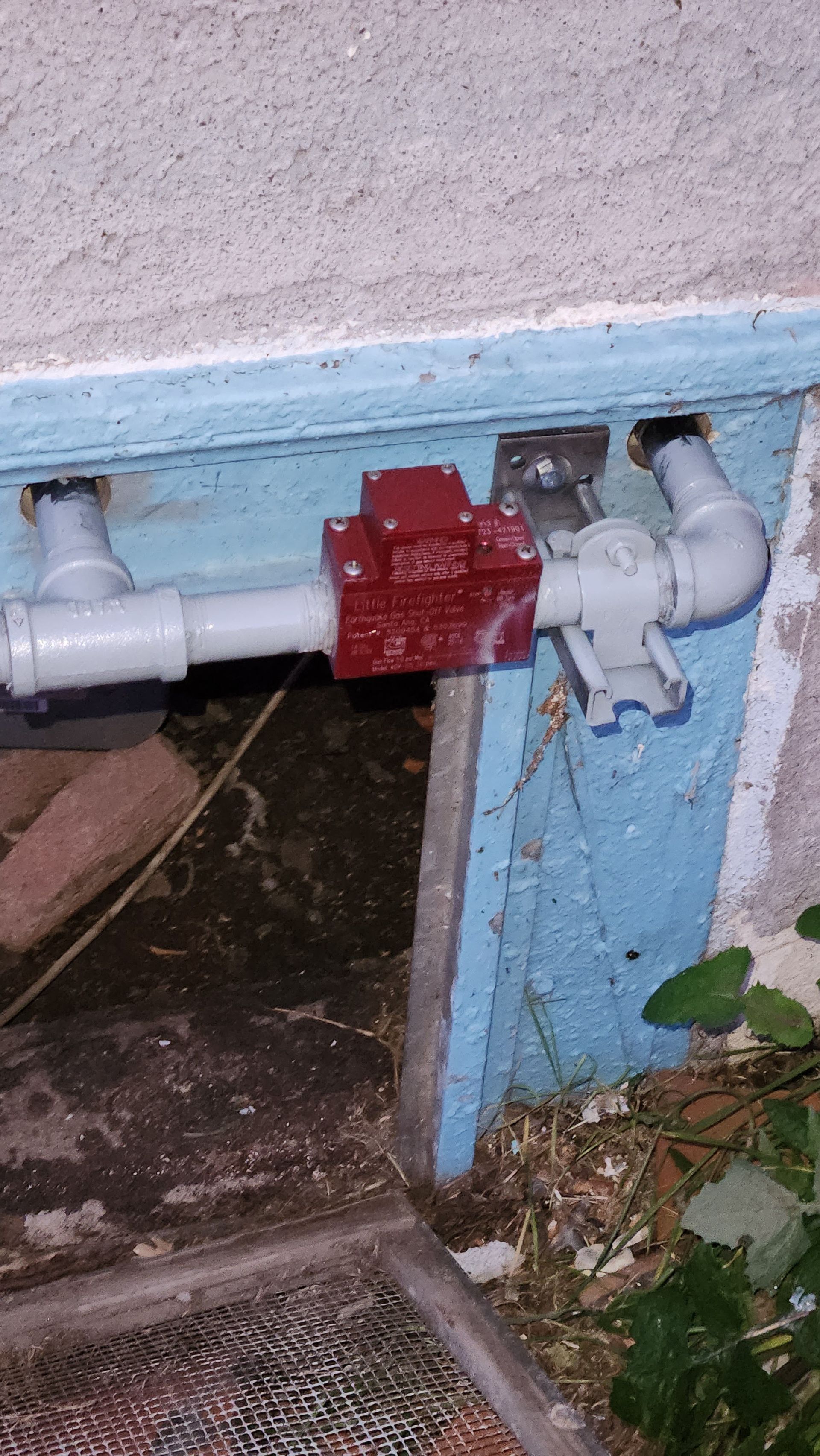 A red valve is attached to a white pipe on the side of a building.