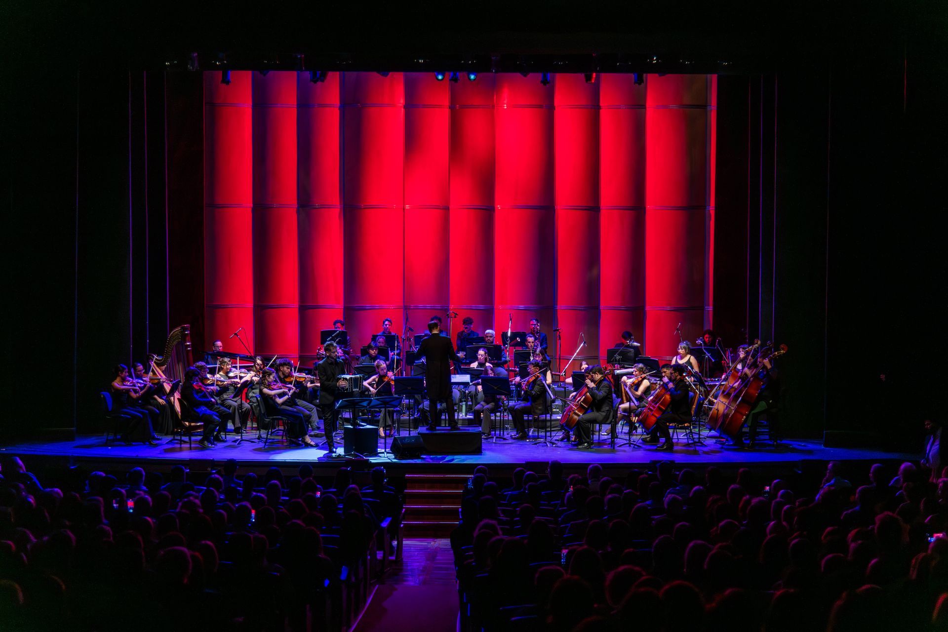 Orquesta en escena, con director, actuando sobre un fondo iluminado con luz roja. El público en primer plano.