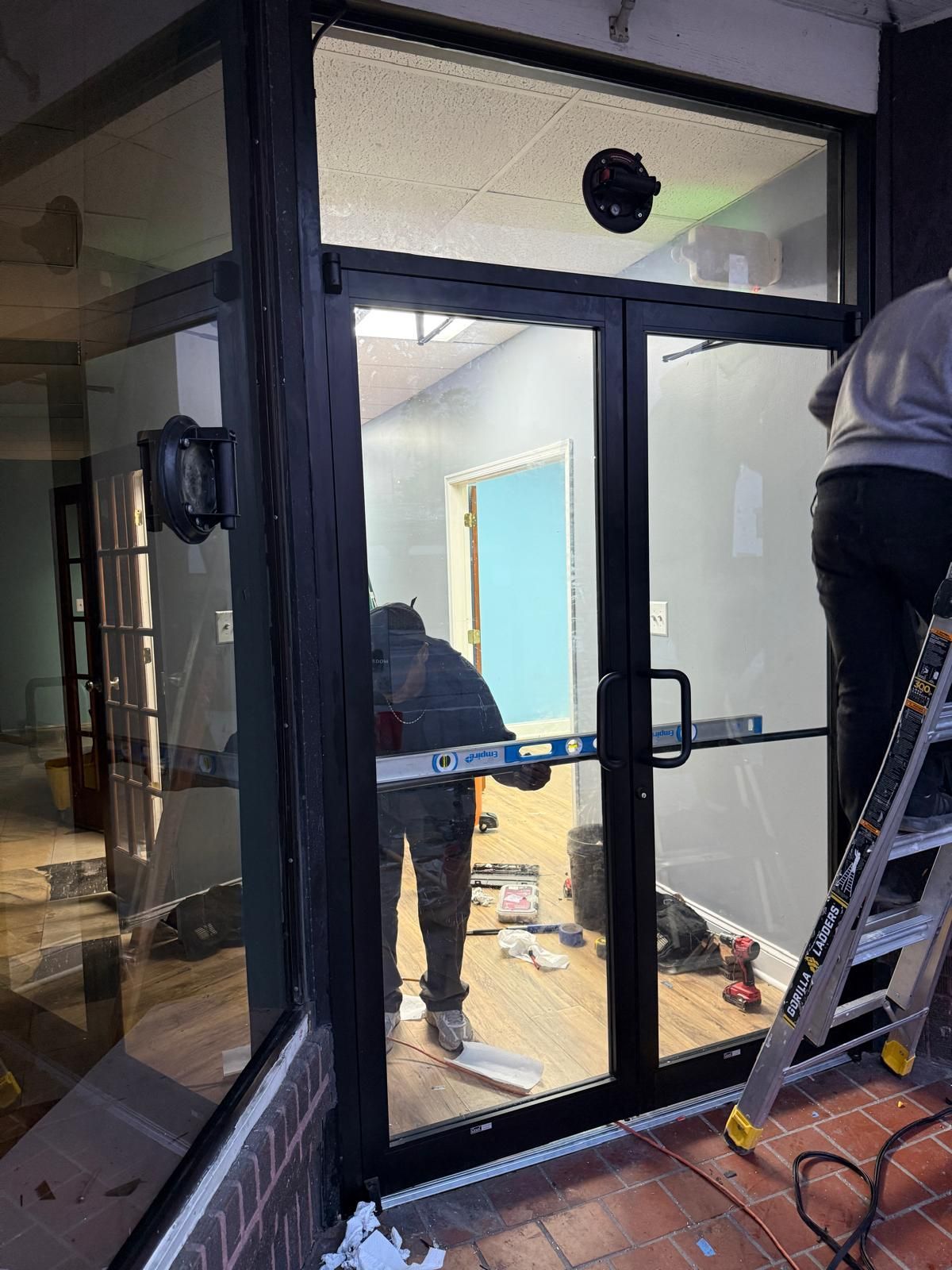 People installing a black-framed glass door, inside a building with a brick exterior.