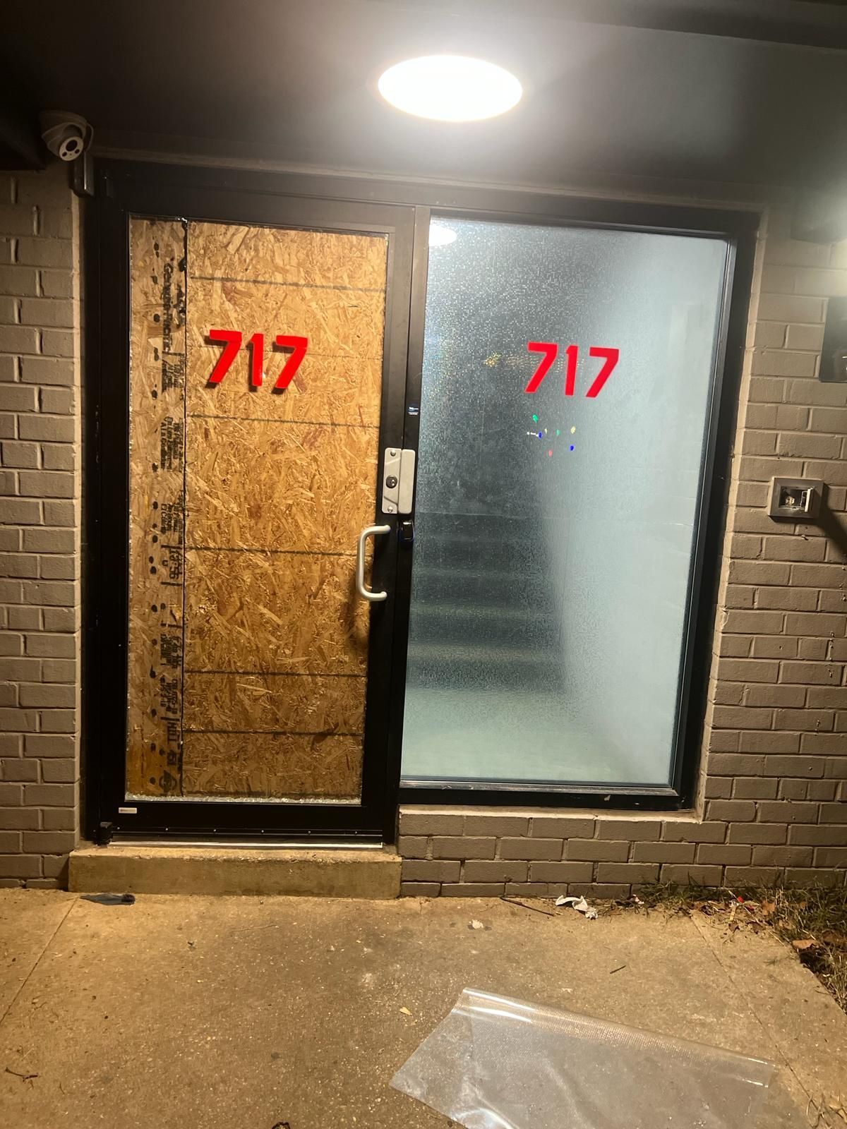 Entrance to building 717, one door boarded up with plywood, the other frosted glass, a security camera above.