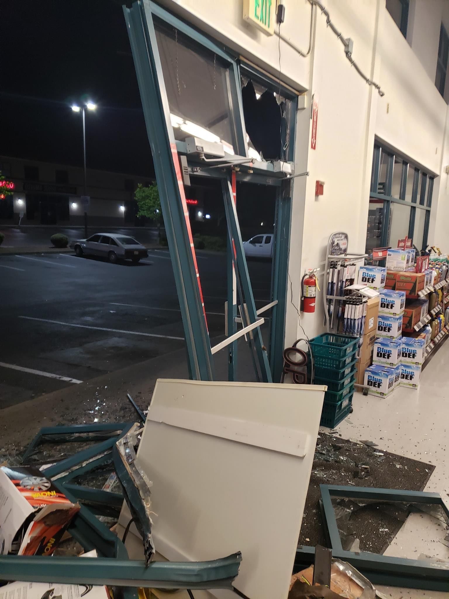 Smashed storefront door with broken glass, debris scattered inside. Exterior nighttime scene with cars.