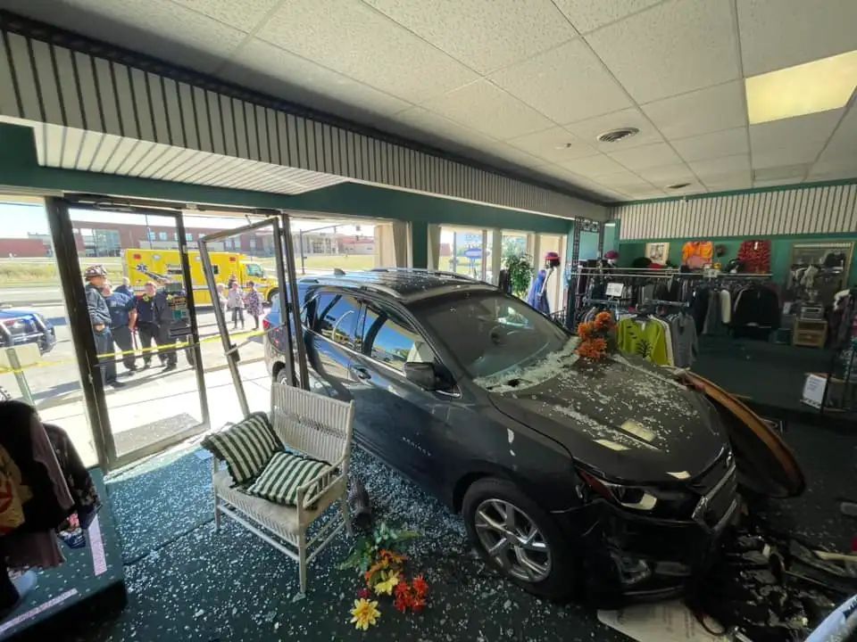 Car crashed inside a store, shattering glass. Emergency responders and onlookers outside.