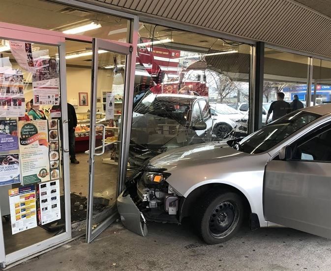 Car crashed into a storefront. Silver car and white truck inside, broken glass, emergency vehicles present.