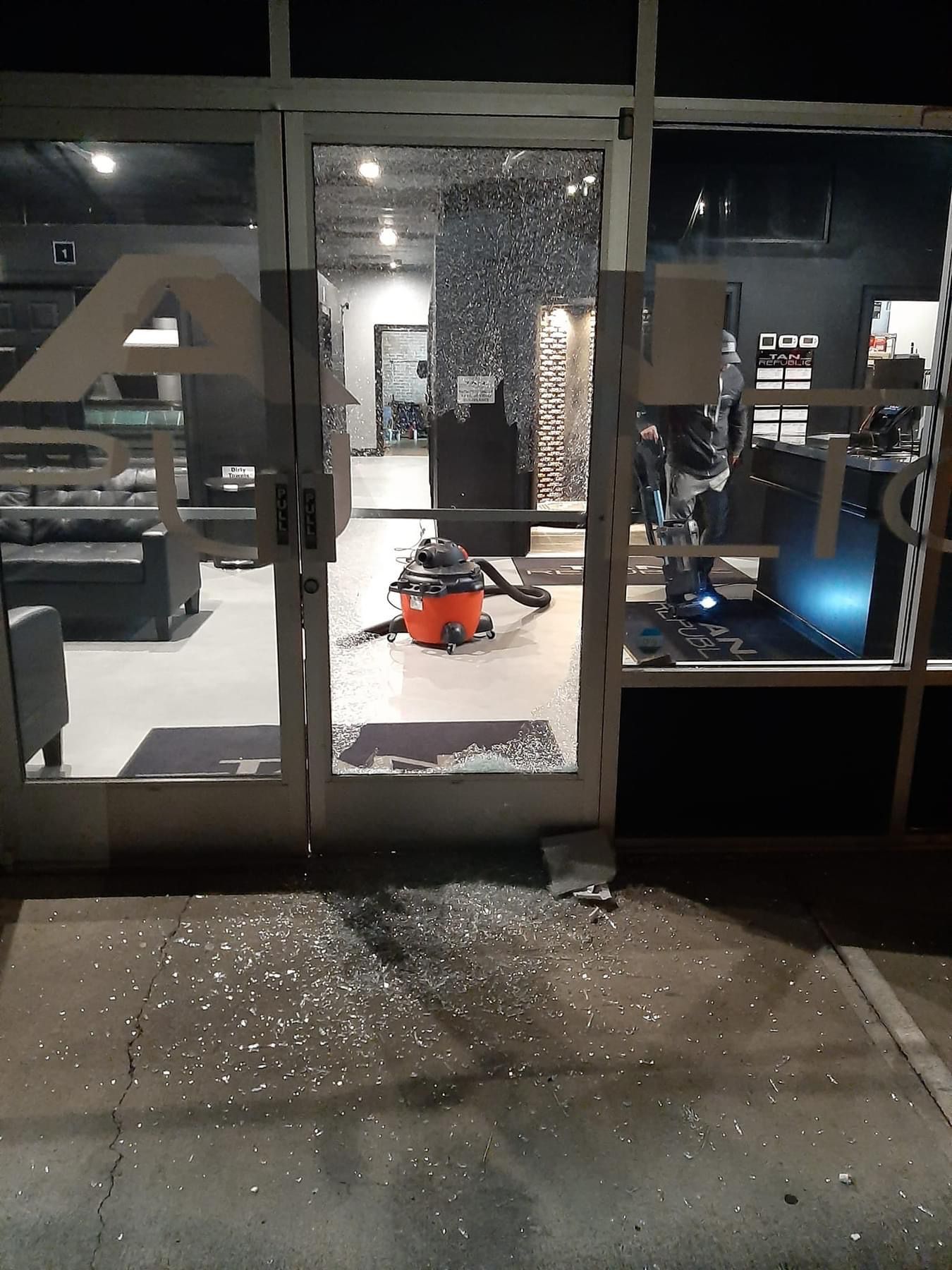 Shattered glass door of a business; vacuum inside, debris on the ground at night.