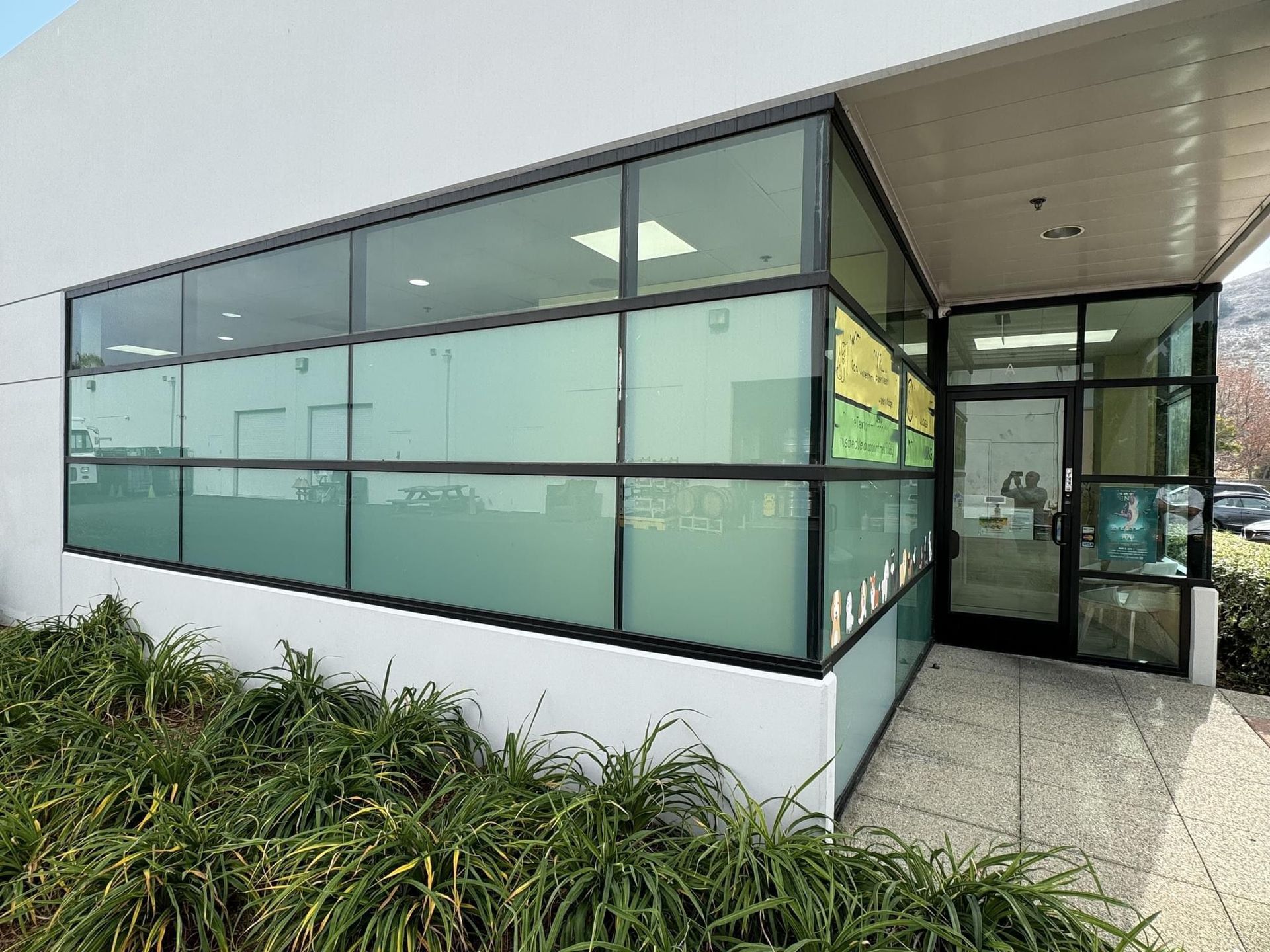 Exterior view of a commercial building with large windows. Frosted glass obscures view, dark frame. Lush green bushes below.