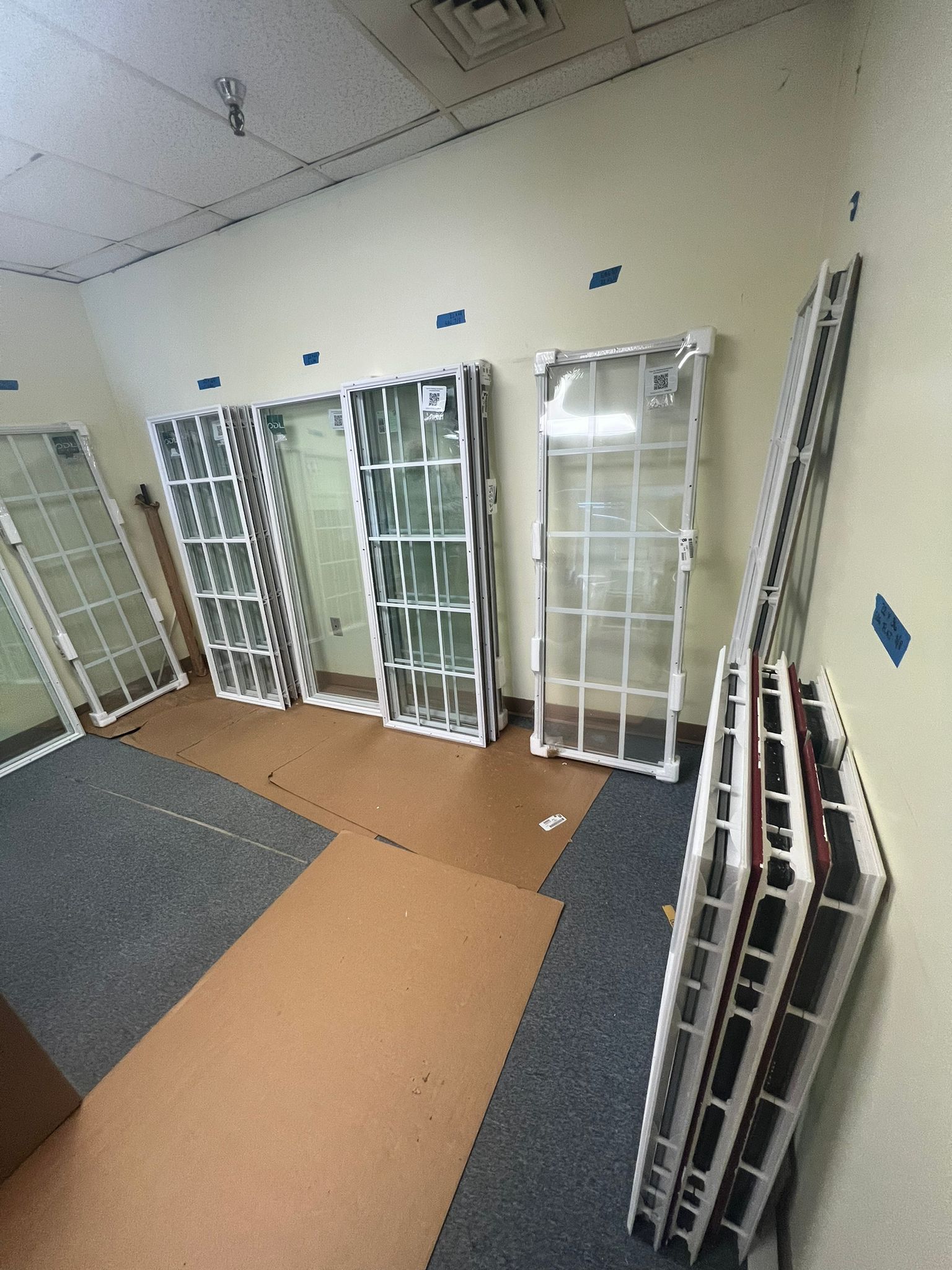 Glass windows with white frames lean against beige walls in a room with a brown floor and blue tape.