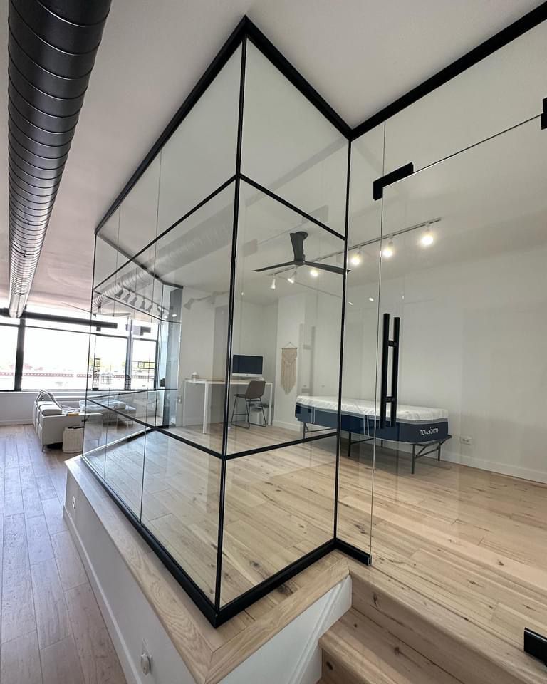 Modern interior with glass-walled room on a raised platform. Black framing, light wood floors, and minimalist furniture.