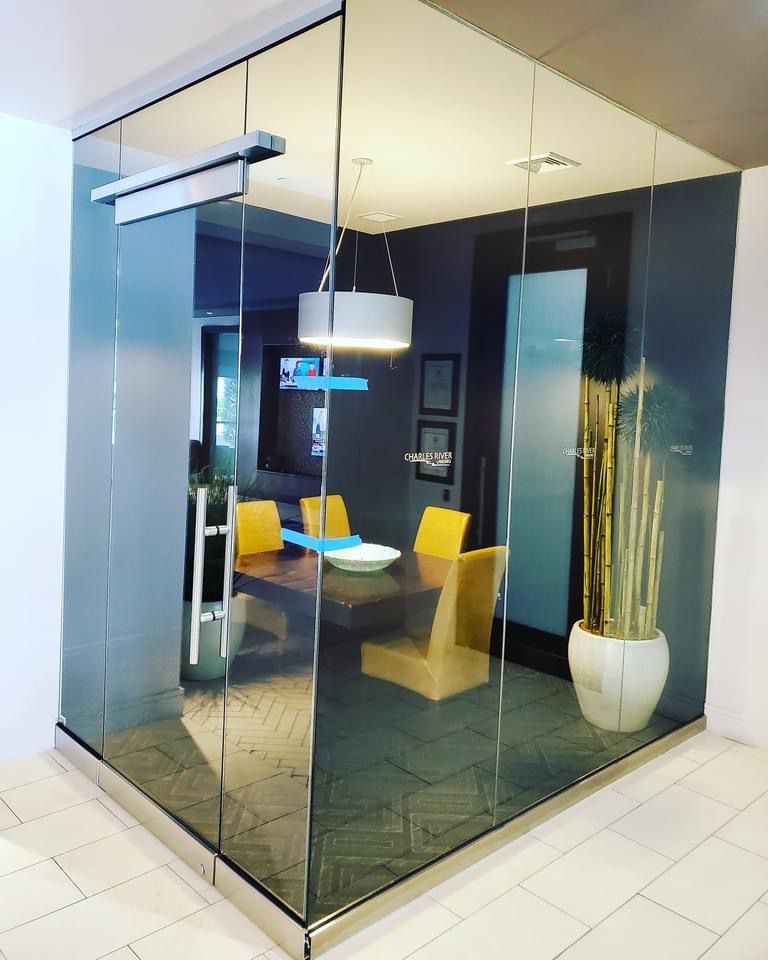 Glass-walled meeting room with table, chairs, and potted plant, located in a hallway.