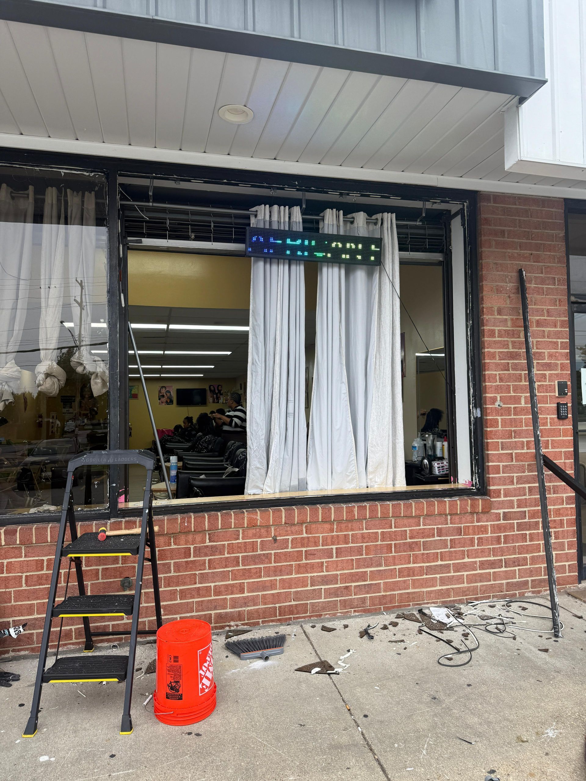 Storefront with a shattered window, brick exterior, and a ladder, bucket, and debris on the ground.