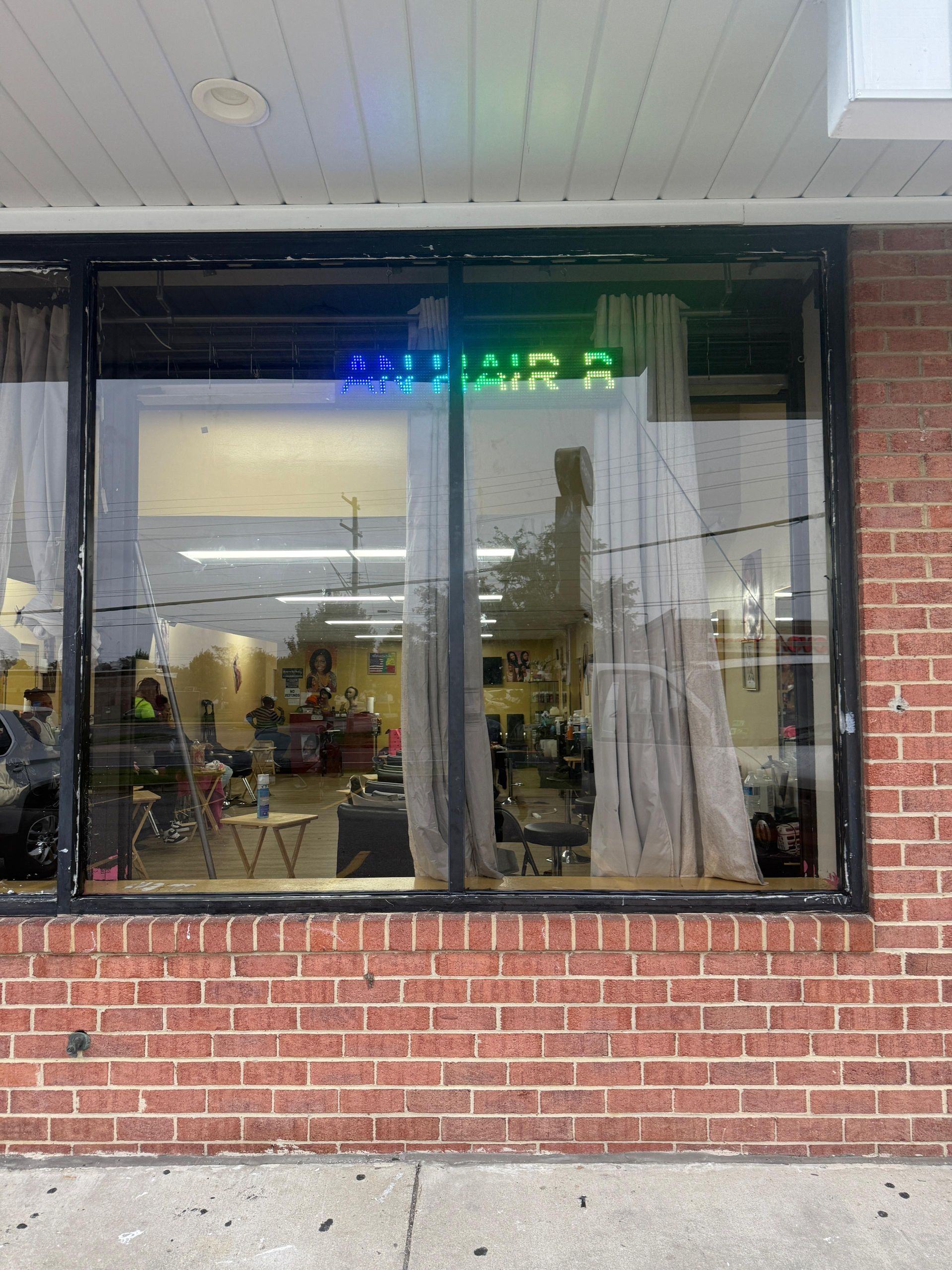 Exterior view of a hair salon with a neon sign in the window, reflected in the glass, and red brick facade.