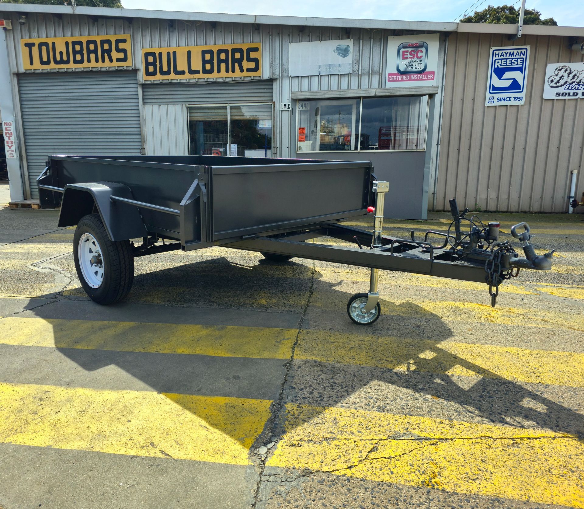 Black utility trailer parked in front of a building with signs for towbars and bullbars.