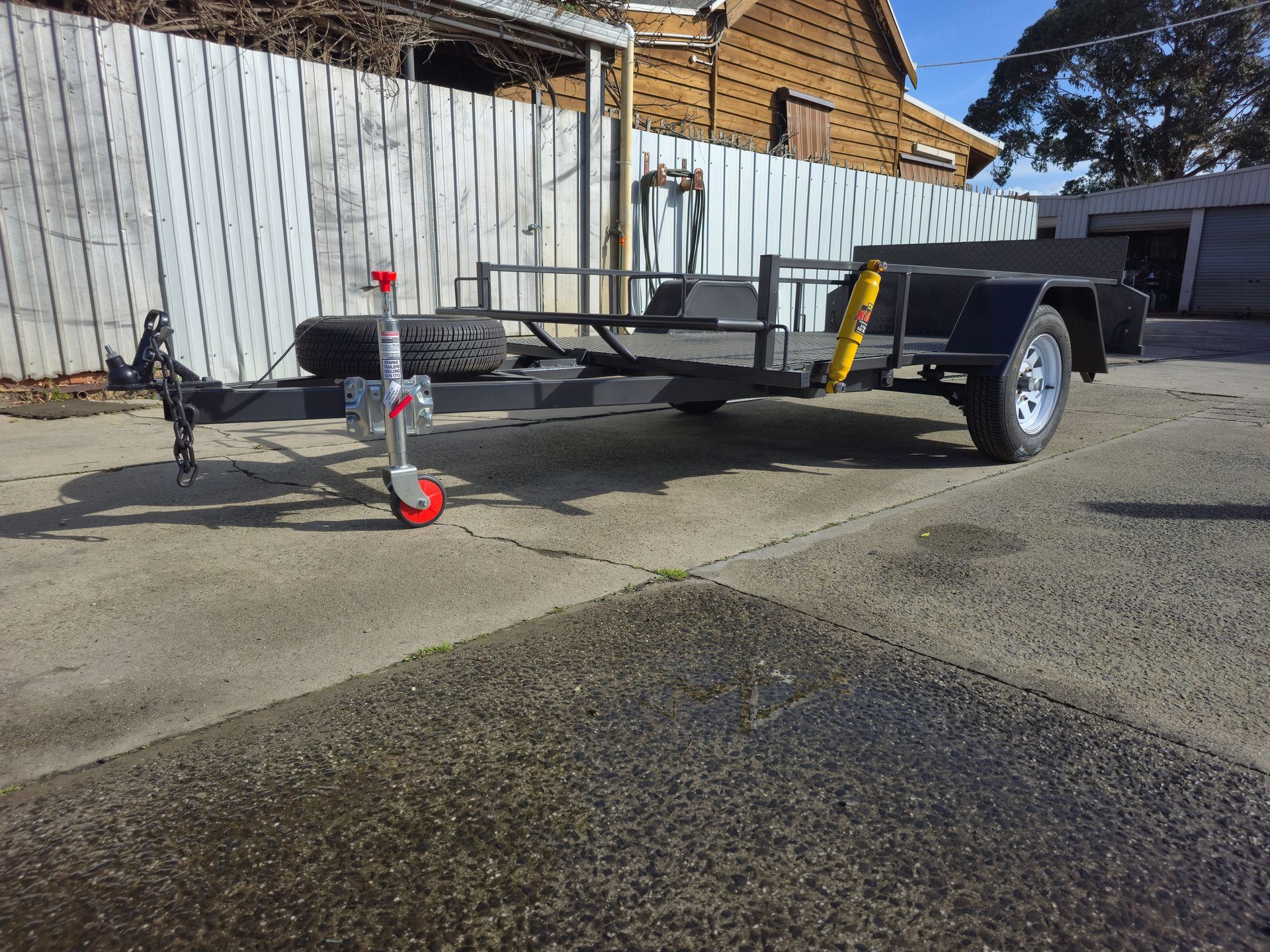 Black utility trailer on a paved surface, spare tire, red reflectors, wooden fence background.