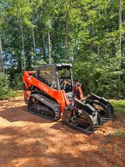 Orange Kubota track skid steer with grapple attachment in a wooded area.