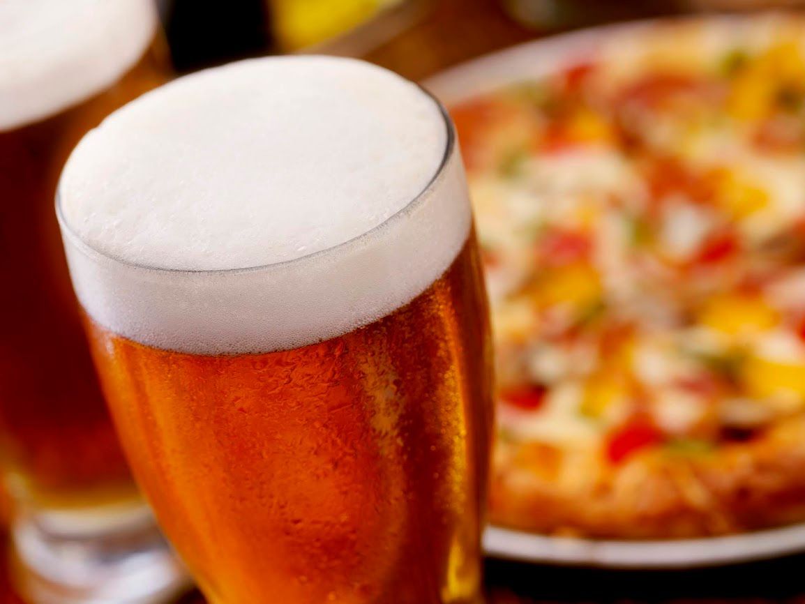 Primo piano di un bicchiere di birra dorata con una spessa schiuma bianca, con una pizza sfocata sullo sfondo.