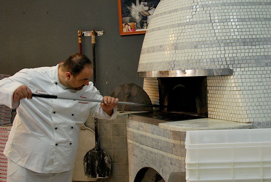 Uno chef in uniforme bianca usa una pala dal manico lungo per cuocere una pizza in un grande forno a cupola rivestito di piastrelle bianche.