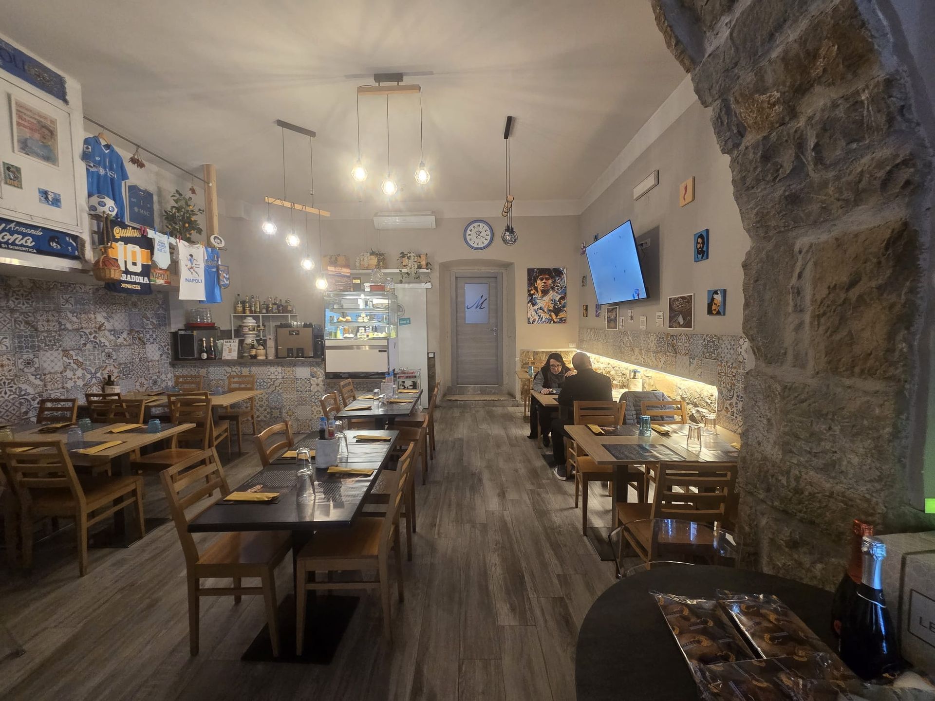 Vista interna di un accogliente ristorante con tavoli e sedie in legno, pareti in pietra, lampade a sospensione e maglie sportive in mostra.