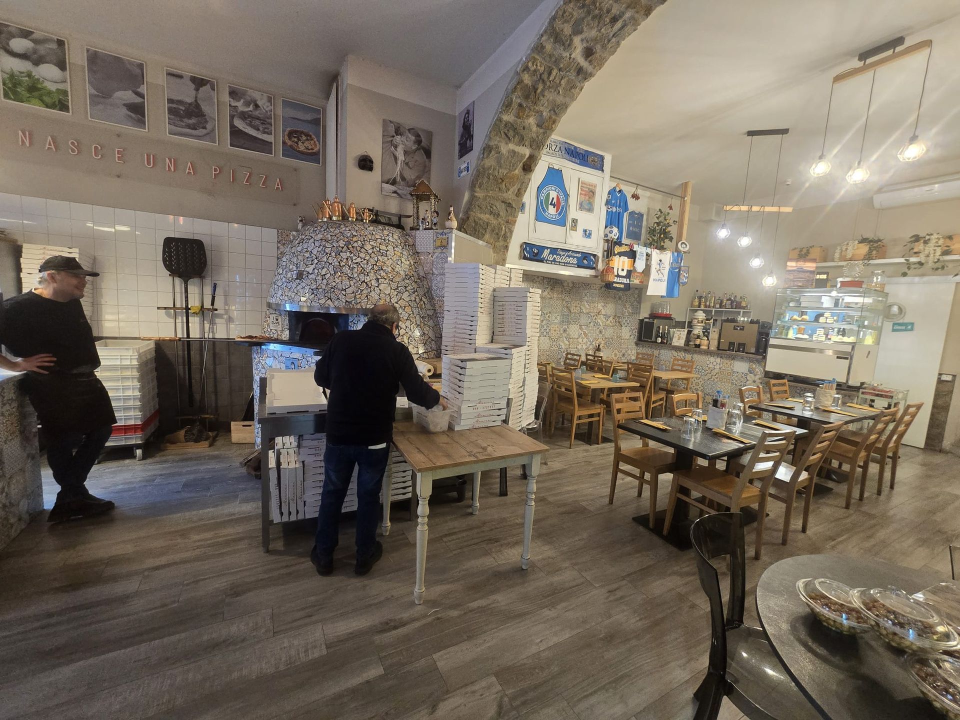 Interno di un ristorante con forno per la pizza, due membri dello staff al lavoro e diversi tavoli in un ambiente rustico.