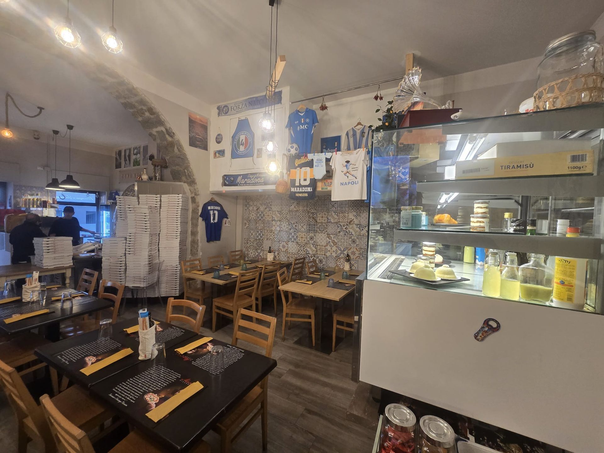 Interno di un accogliente ristorante con tavoli in legno, pareti piastrellate e una vetrina piena di bevande e prodotti alimentari.