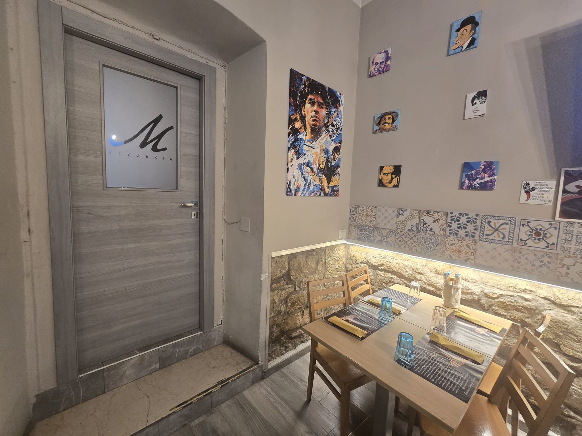 Un accogliente angolo pranzo con tavolo e sedie in legno e una parete in pietra, decorata con poster e una porta con il logo della lettera M.
