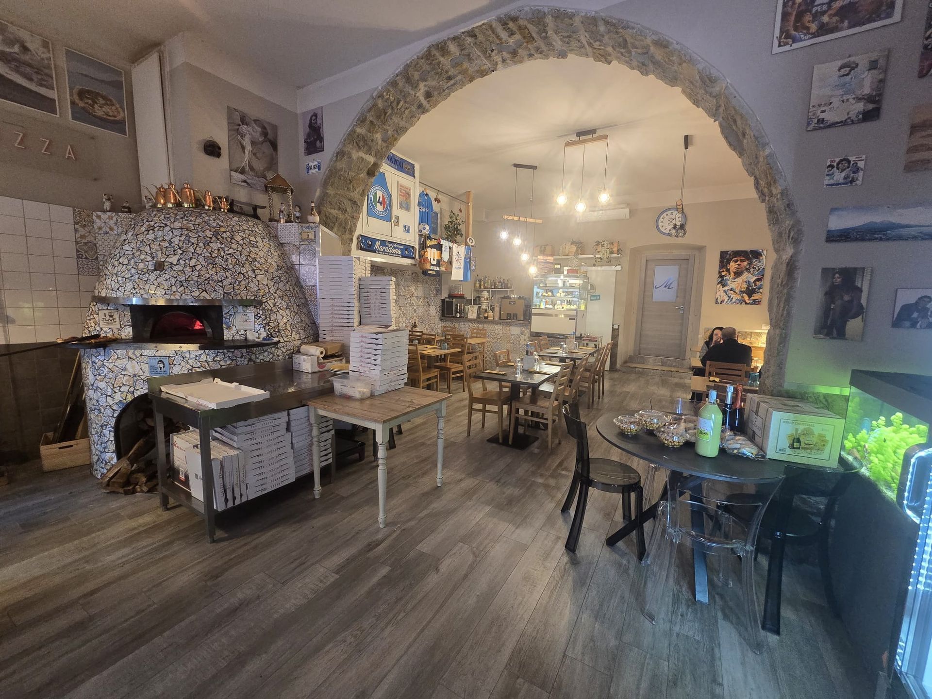 L'interno accogliente di un ristorante è caratterizzato da un arco in pietra, un forno per la pizza rivestito in piastrelle, tavoli in legno e una calda illuminazione dall'alto.