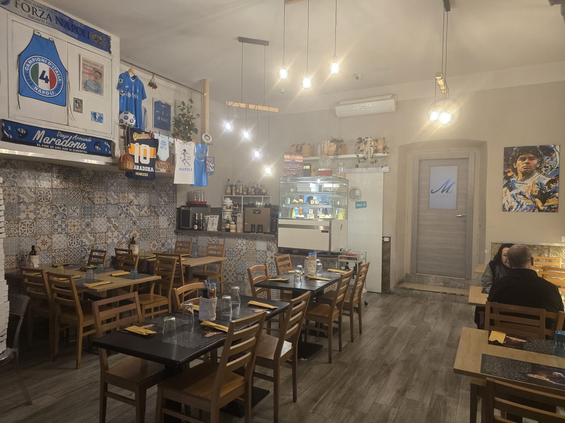 L'interno di un ristorante con tavoli e sedie in legno, decorato con maglie da calcio e opere d'arte che celebrano Diego Maradona.