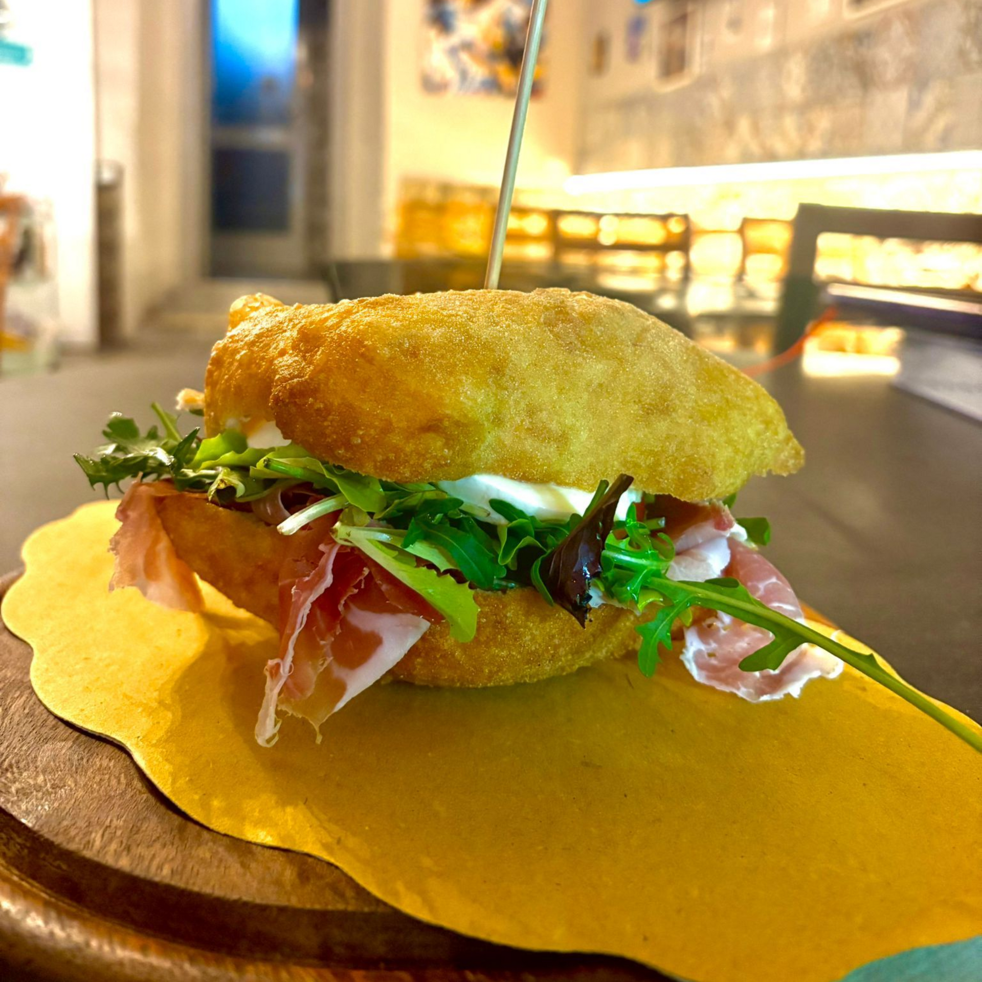 Un panino fritto dorato, farcito con prosciutto crudo, verdure fresche e formaggio morbido, servito su un tagliere di legno.