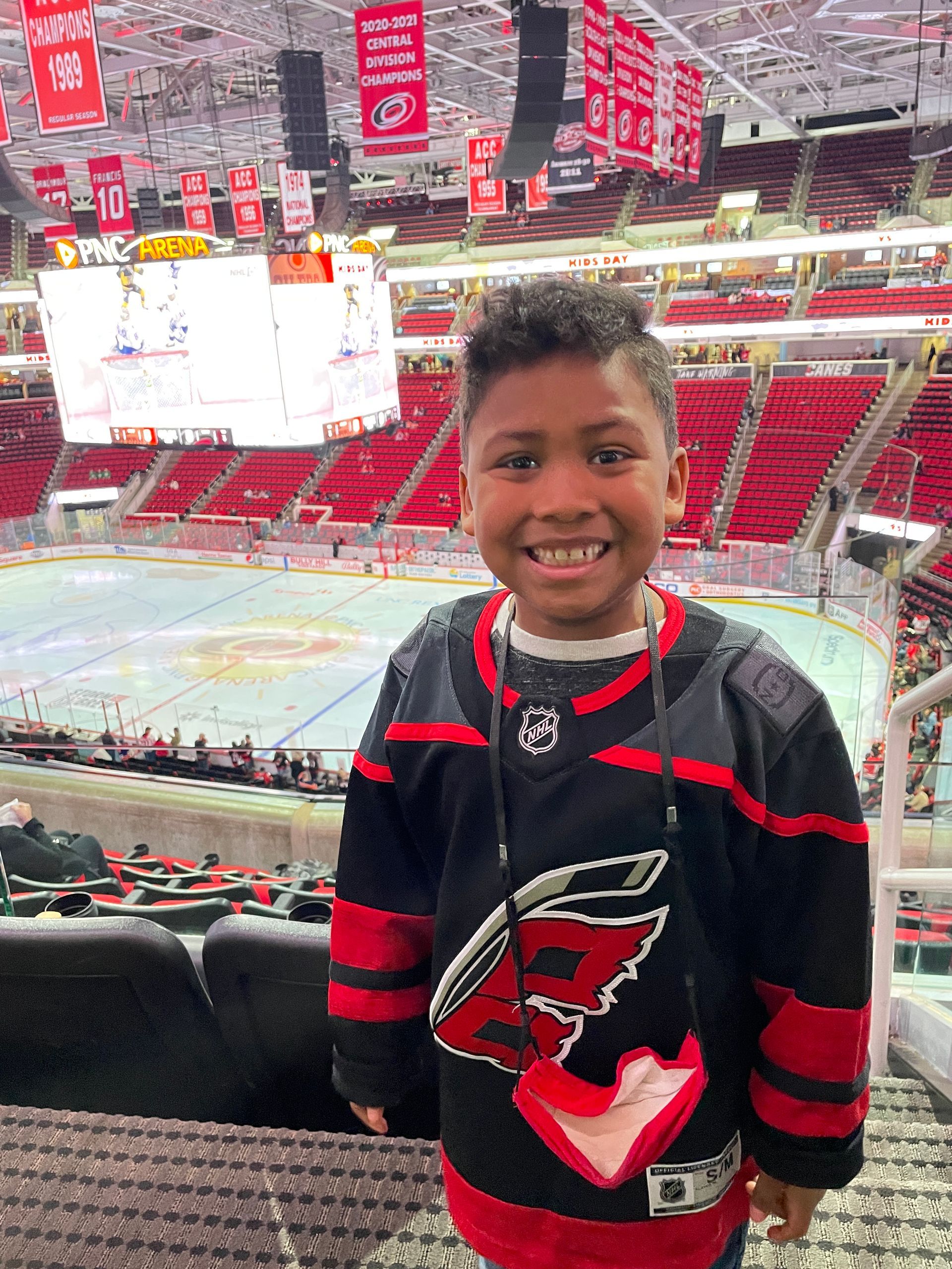 A young boy in a hockey jersey is standing in front of a hockey rink.