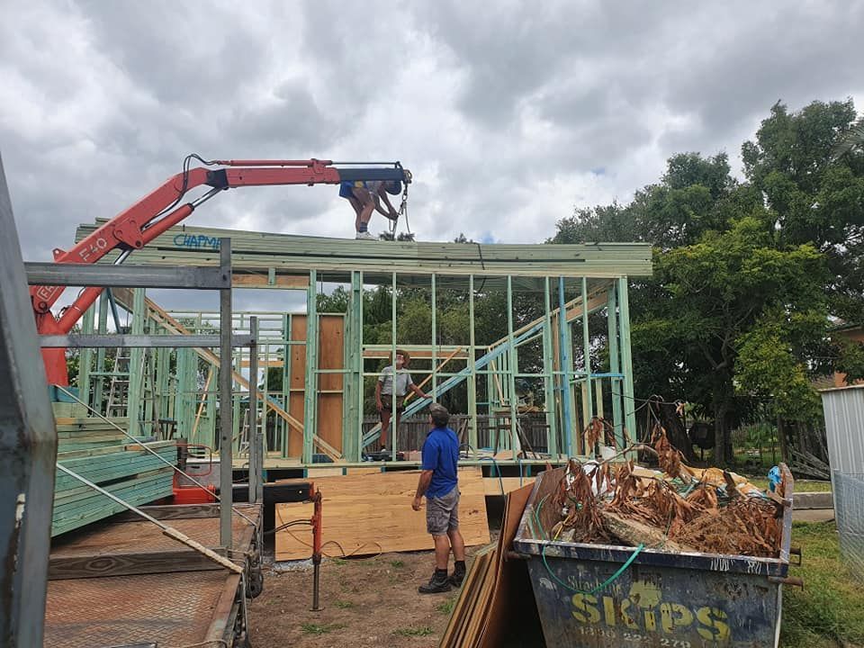 Construction of a wood-framed building. A crane lifts materials onto the roof while a worker observes near a debris bin. Cloudy sky.