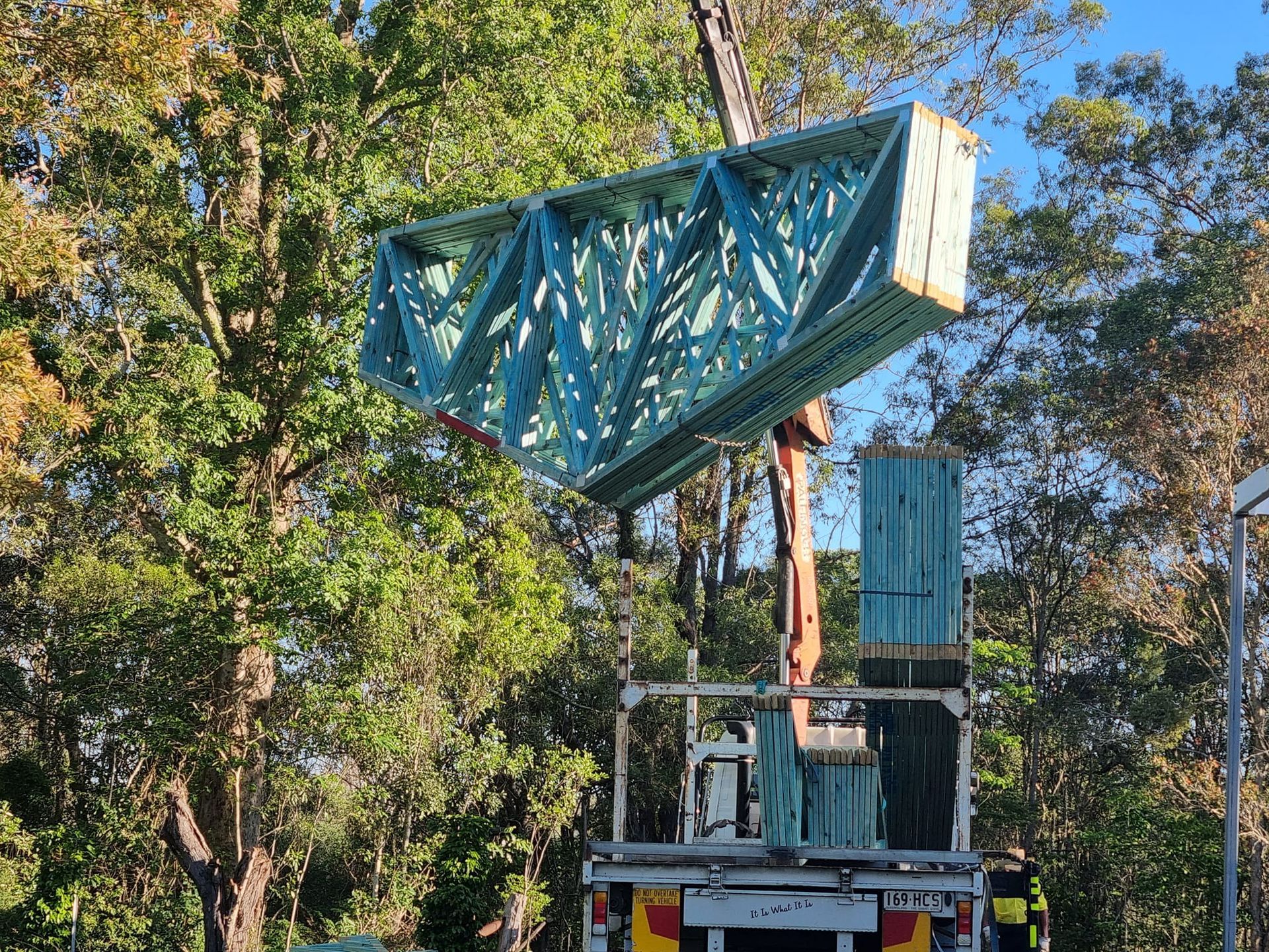 A crane lifts a large wooden truss over a truck bed, near trees and a road.