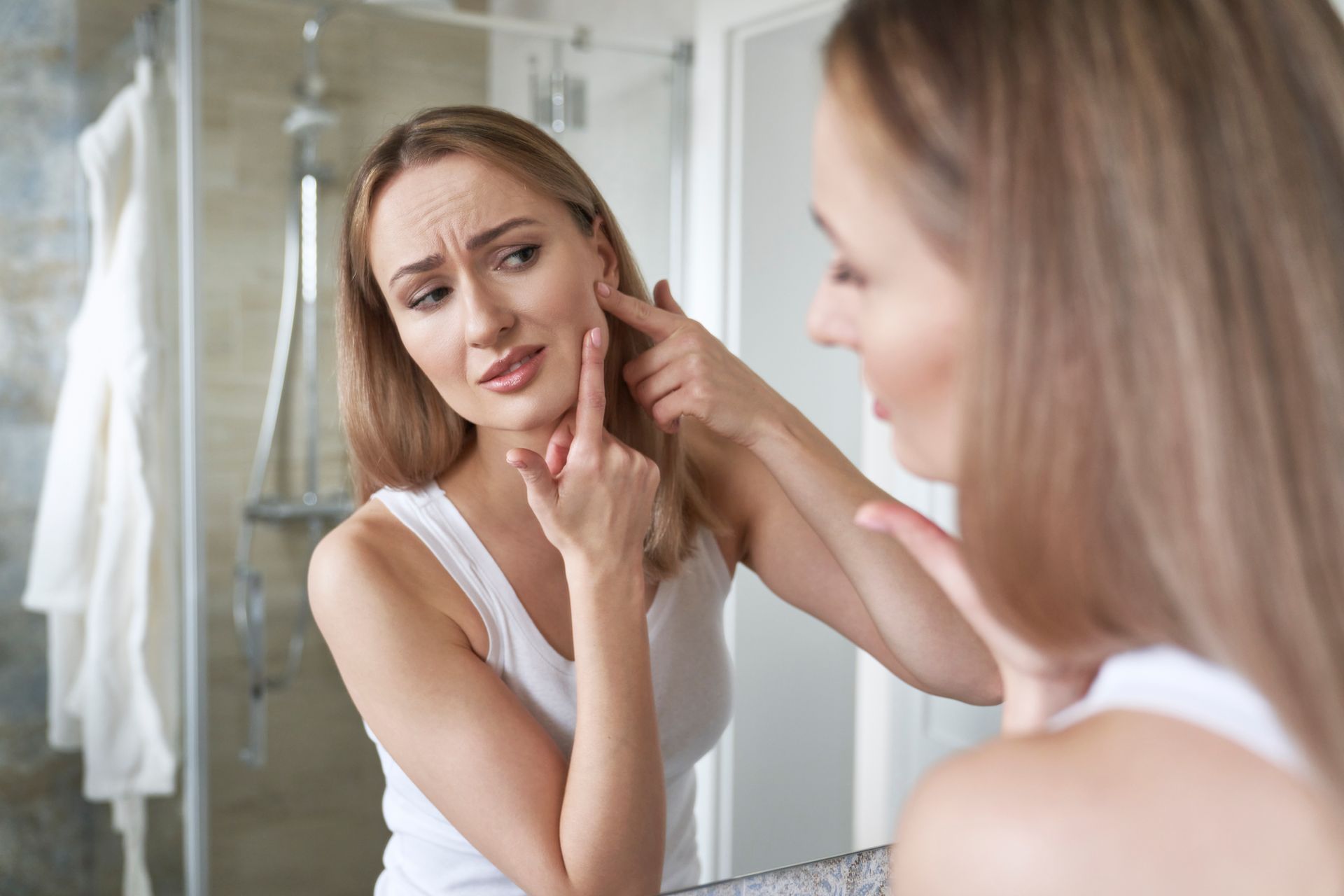 Woman in bathroom looks in mirror, examining skin on her face.