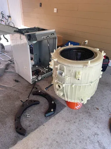 Washing machine disassembled outdoors. Drum and other parts laid out on concrete. White, black, and orange colors.