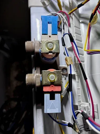 Two water inlet valves in a washing machine, blue and red connectors, attached to the machine's body.