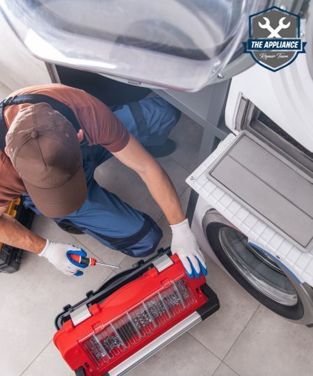 Repair technician fixing a washing machine, using a toolbox and screwdriver, in a laundry room.