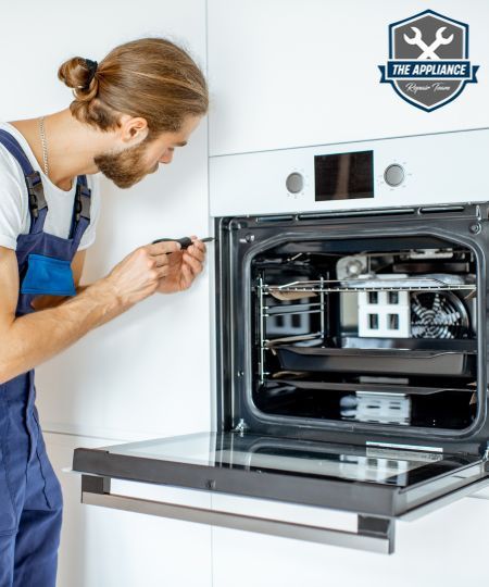 Man in blue overalls repairs an oven with a screwdriver in a kitchen.