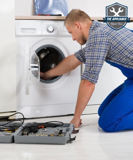 A repairman kneels to fix a washing machine, tools in a case nearby.