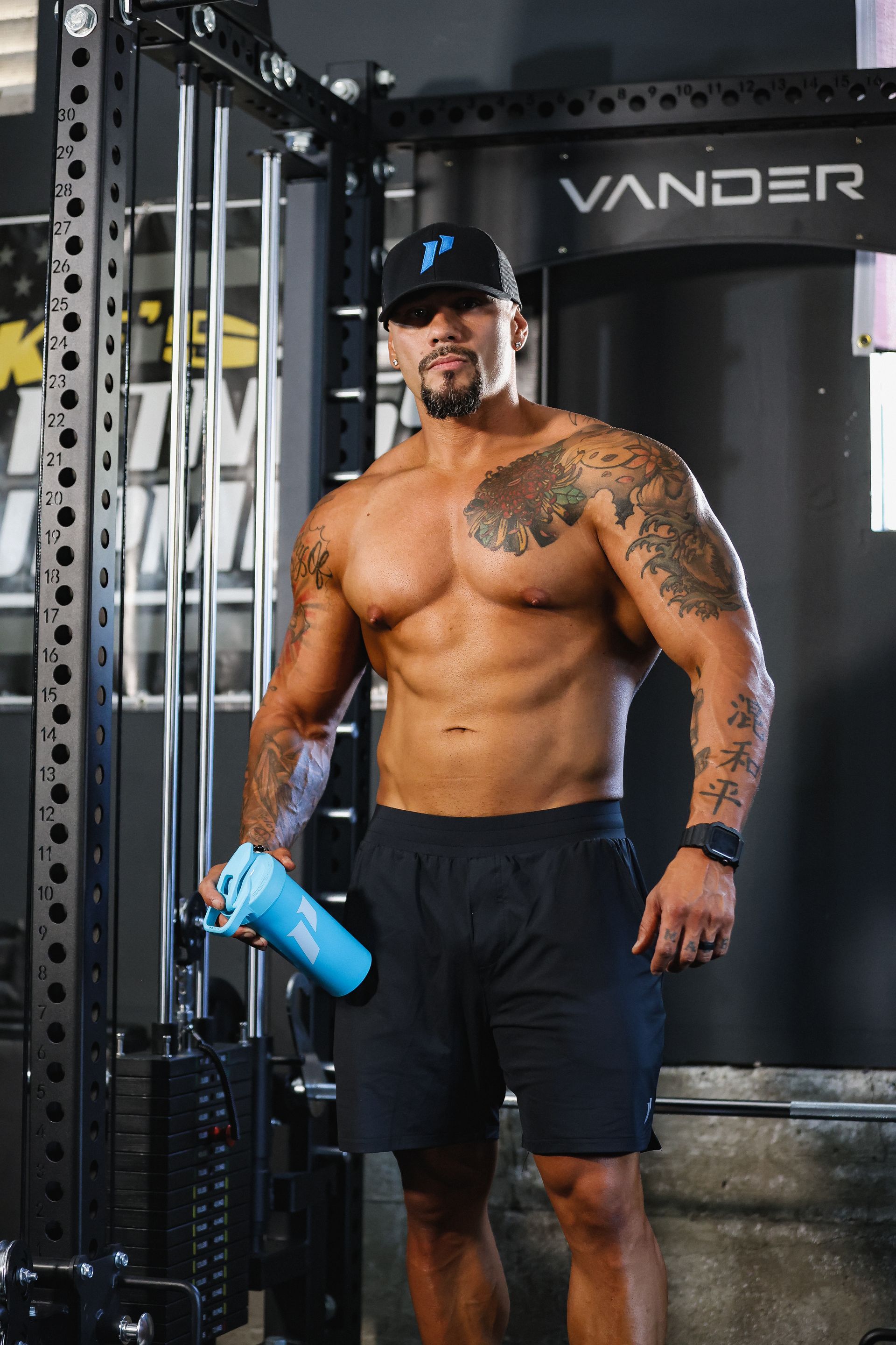 A shirtless man is standing in a gym holding a bottle of water.