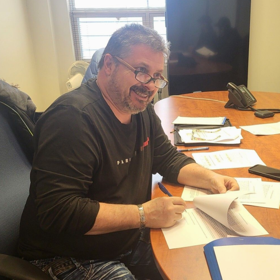 Un homme portant des lunettes signe des documents à une table ronde, en souriant.