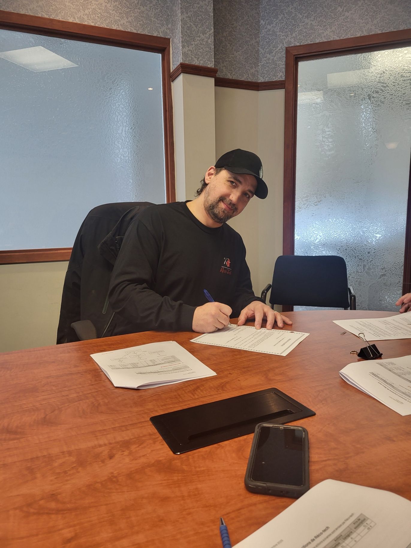 Un homme en chemise et casquette noires est assis à une table, écrivant sur du papier dans une salle de conférence.