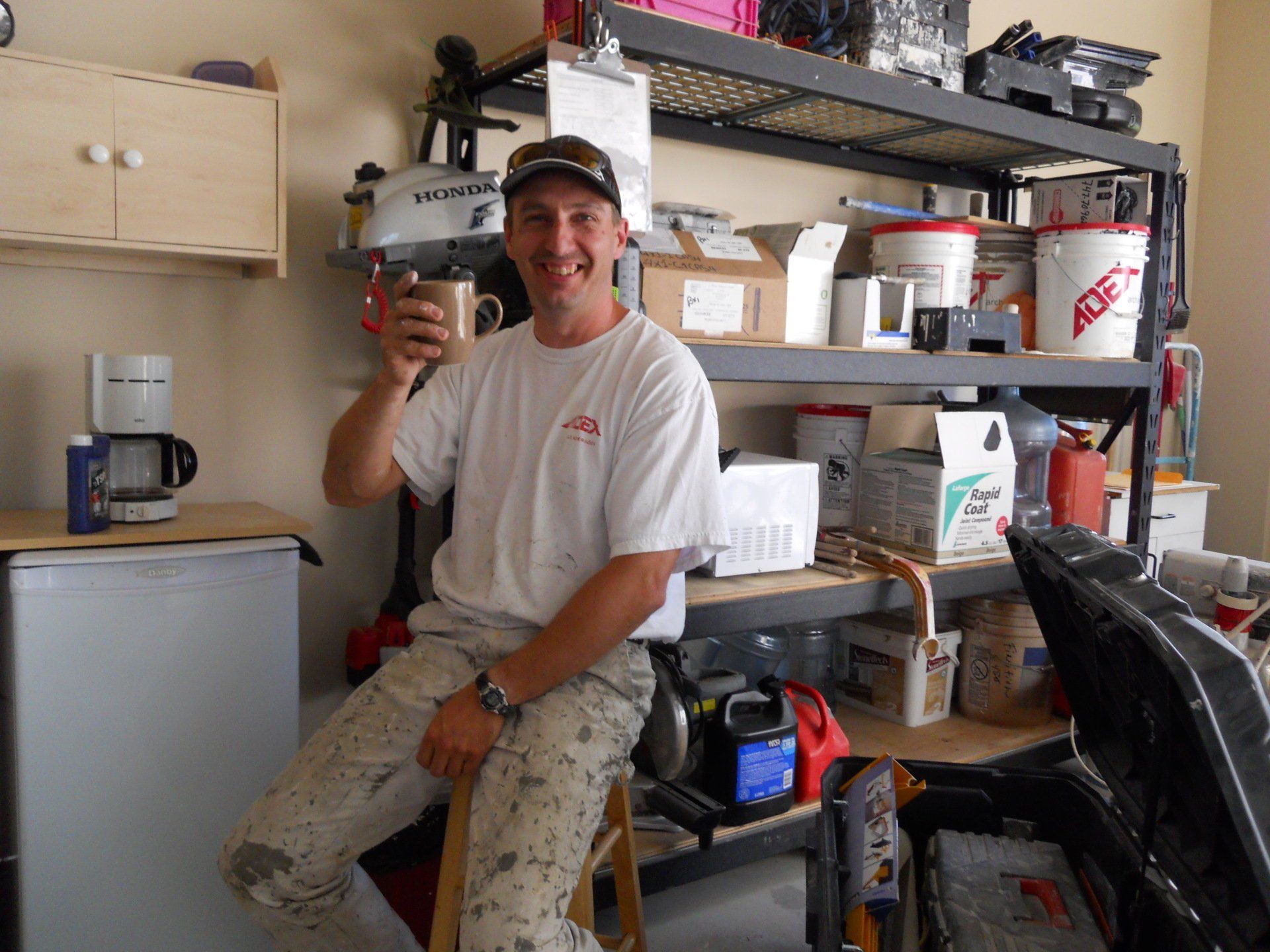 Un homme aux vêtements tachés de peinture sourit, une tasse à la main. Il se trouve dans un garage dont les étagères sont remplies de matériel.