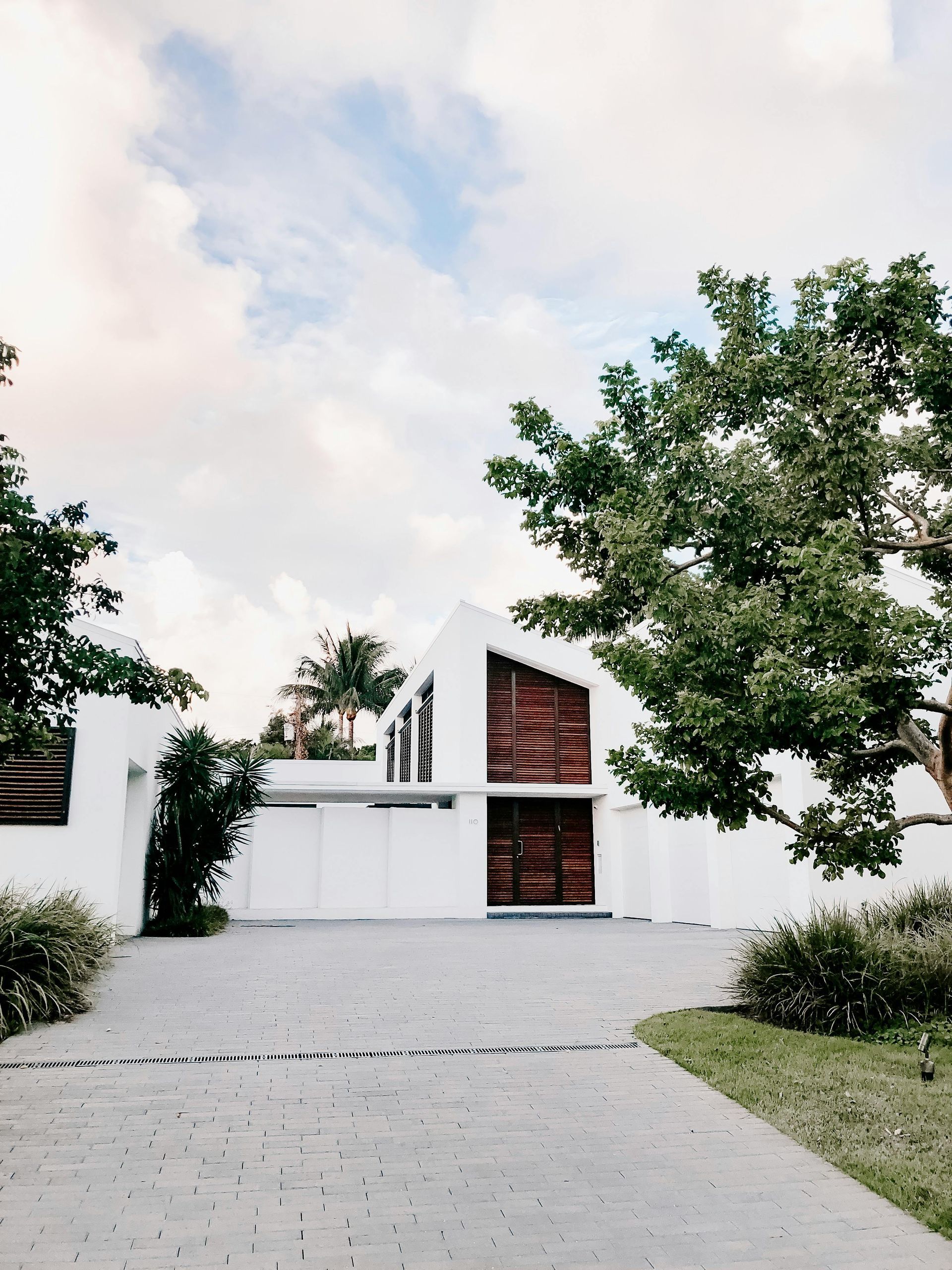 White modern house with wood accents, surrounded by trees and a stone driveway under a cloudy sky.