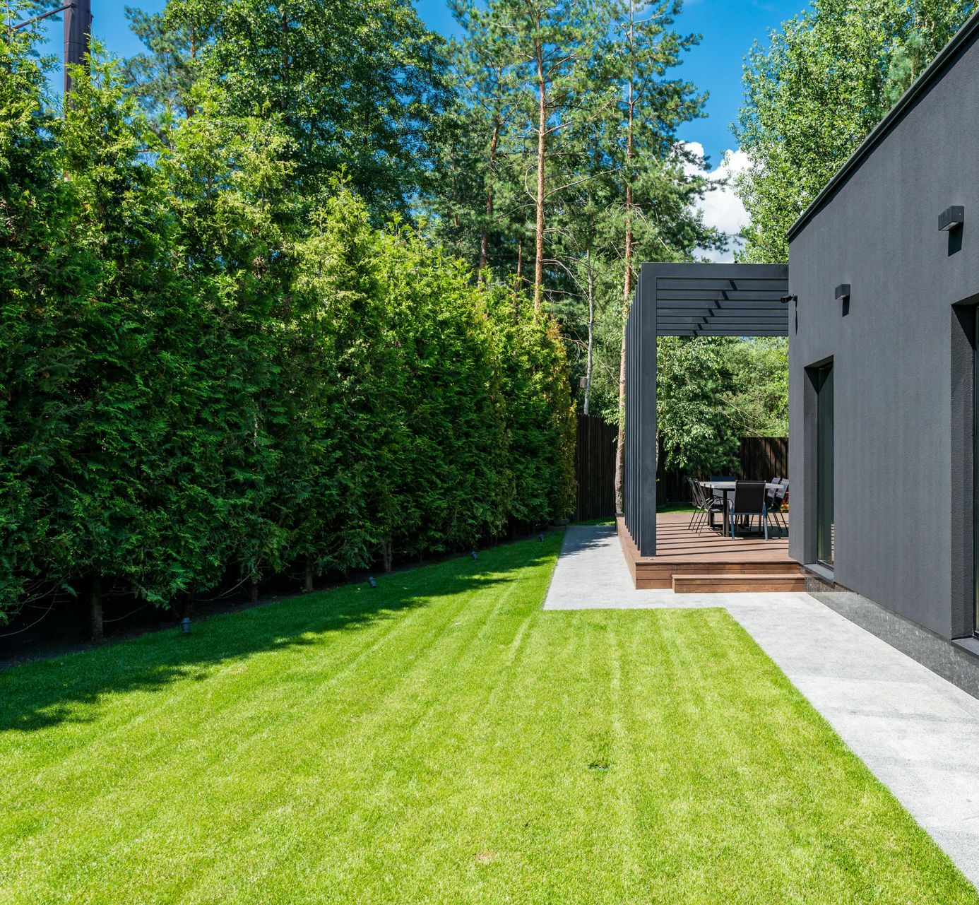 Lush green lawn next to a house with dark gray siding and wooden deck shaded by a pergola, bordered by a tall hedge.