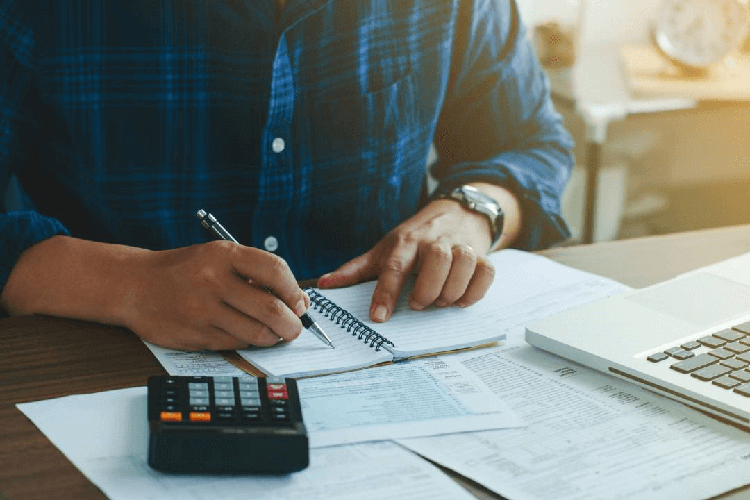 A man is sitting at a desk using a calculator and writing in a notebook.