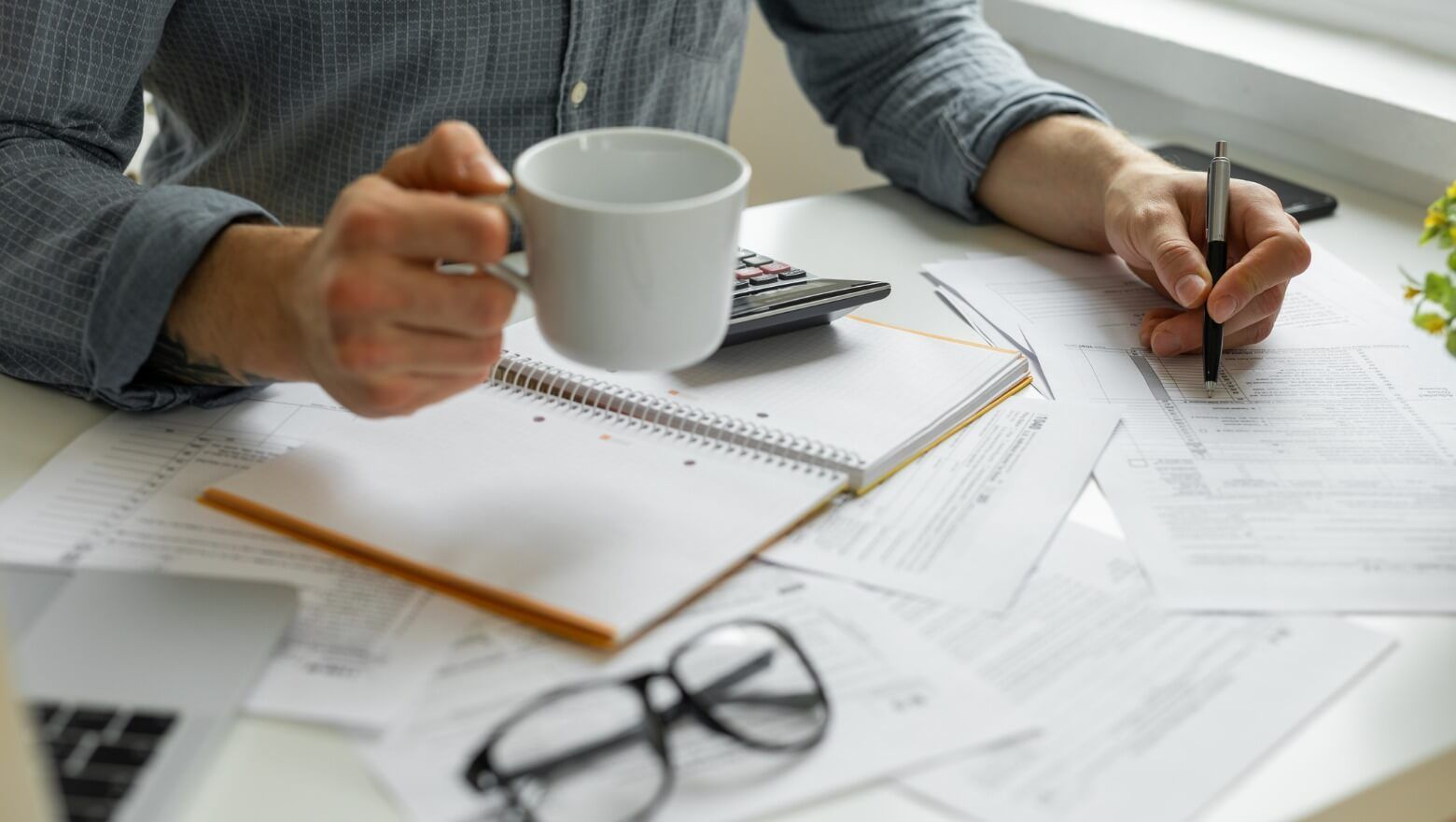 A man is sitting at a desk holding a cup of coffee and writing in a notebook.