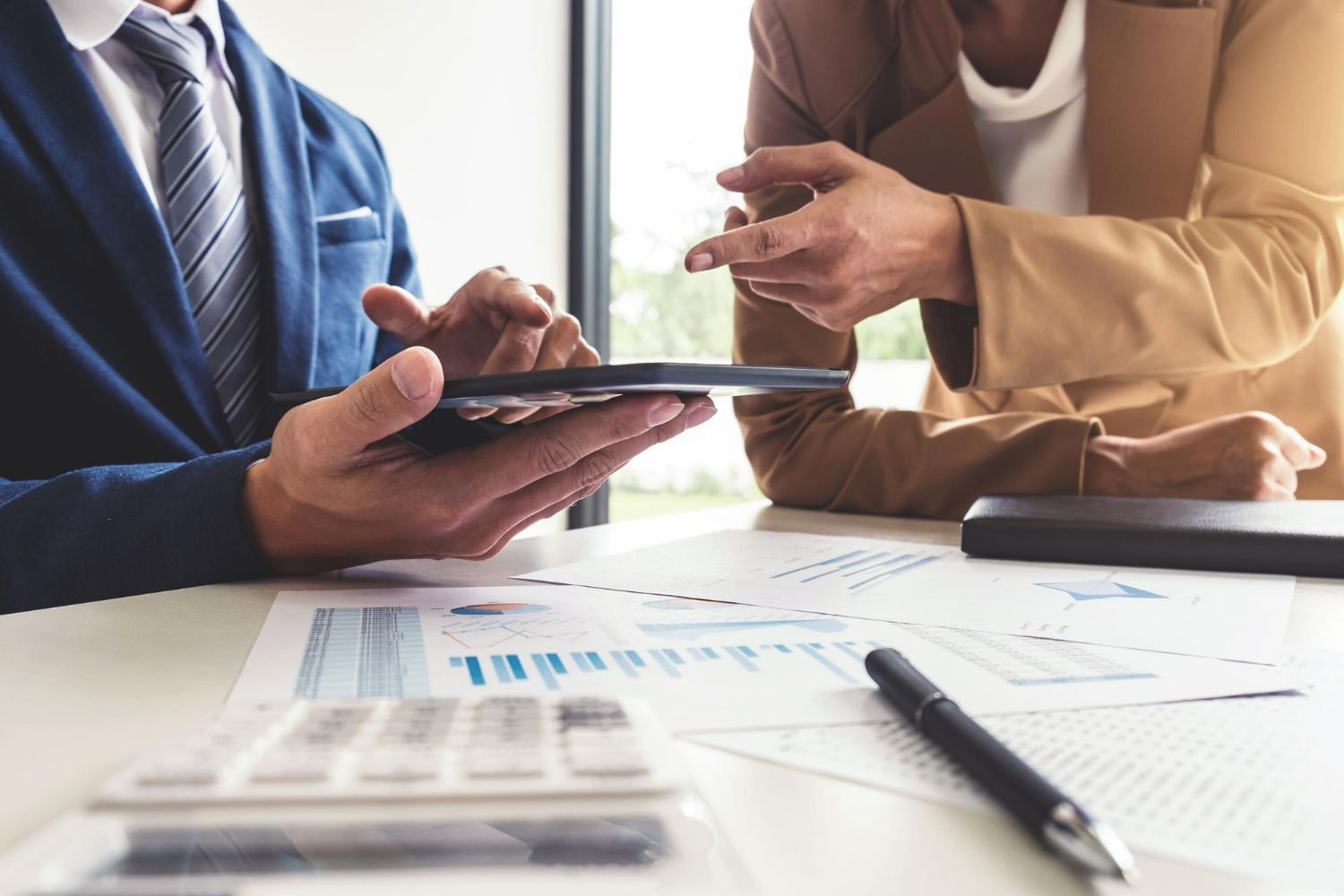 Two businesspeople reviewing financial data on a tablet and papers at a desk.