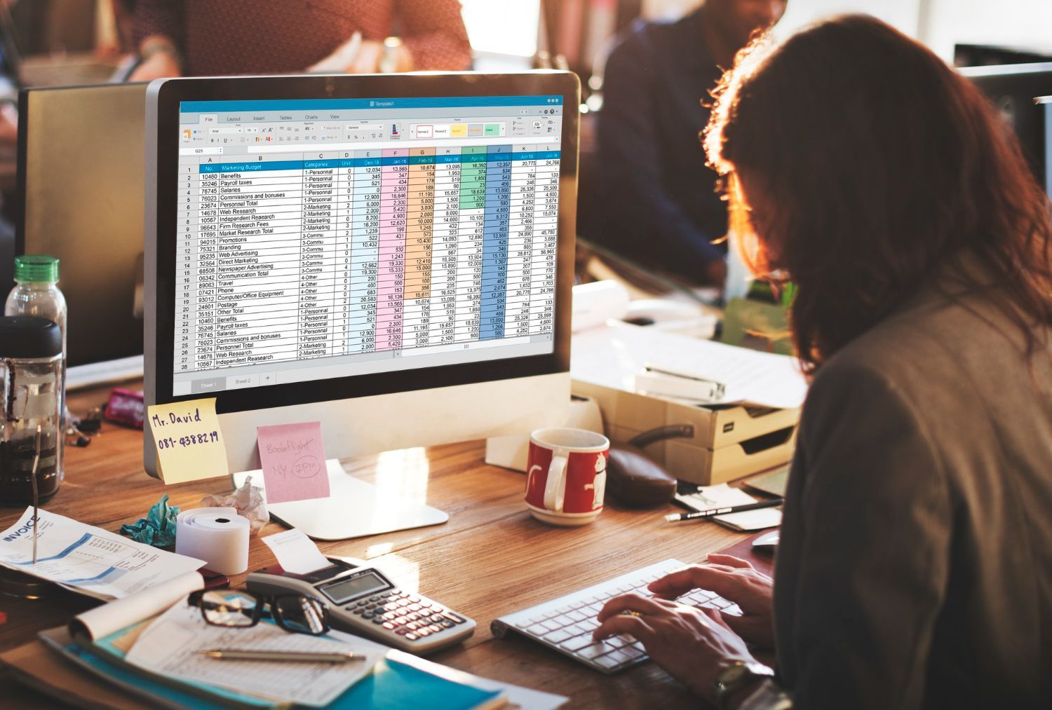 Woman working on a spreadsheet on a desktop computer in an office, typing at keyboard, desk cluttered.